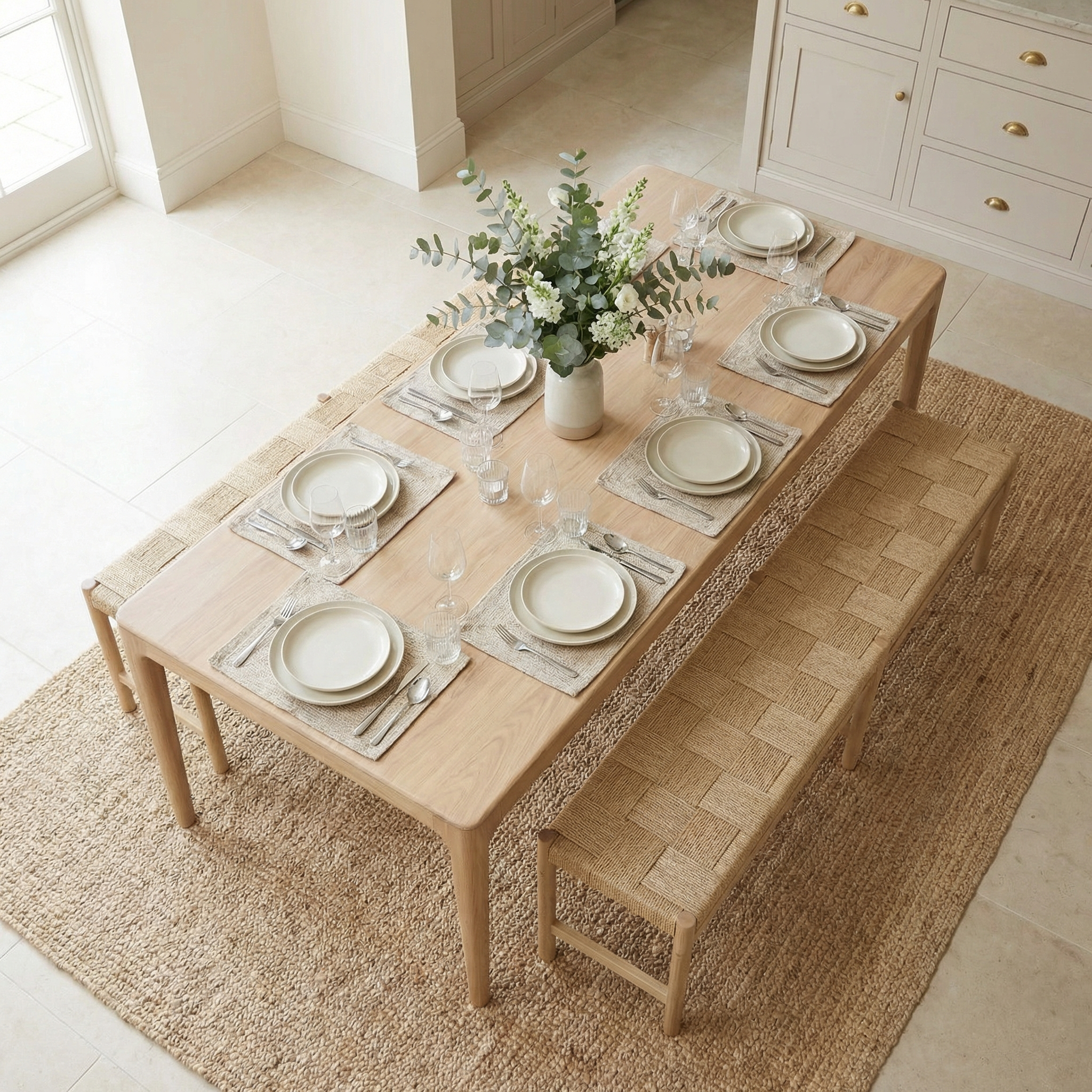 The Marlow 8 Seat Dining Set with Woven Benches in Light Oak is styled with neutral tableware, a vase of flowers, and arranged on a rug.