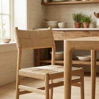 A Marlow Set of 2 Woven Dining Chair in Light Oak is placed beside a matching table in a cozy kitchen decorated with shelves and potted plants.