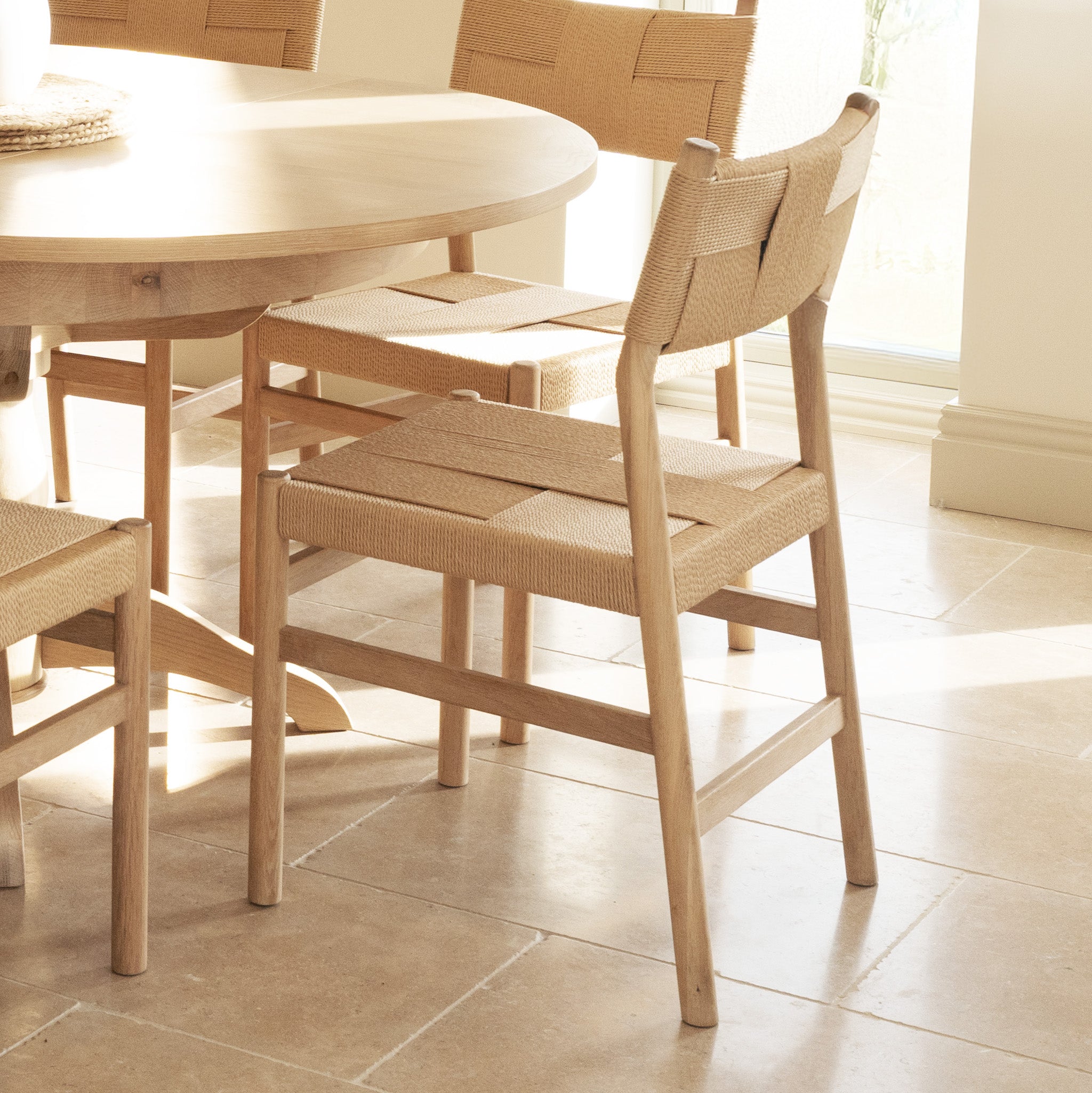 Two Marlow Sets of 2 Woven Dining Chairs in Light Oak with Danish cord seats surround a round wooden table on a tiled floor in a bright room.