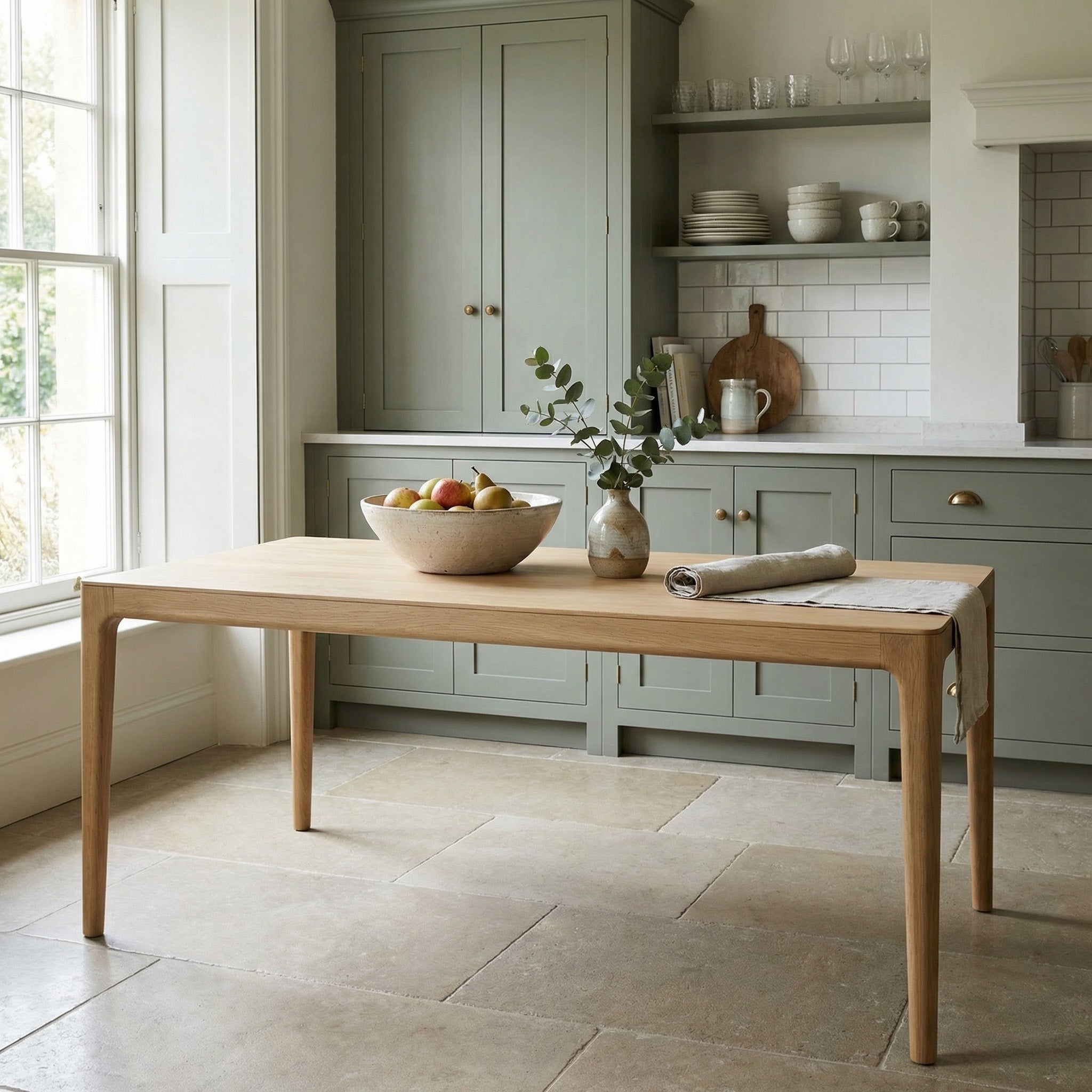 The Marlow 8 Seat Dining Table in Light Oak sits in a kitchen with a fruit bowl, leafy vase, and folded towel, all bathed in soft natural light.