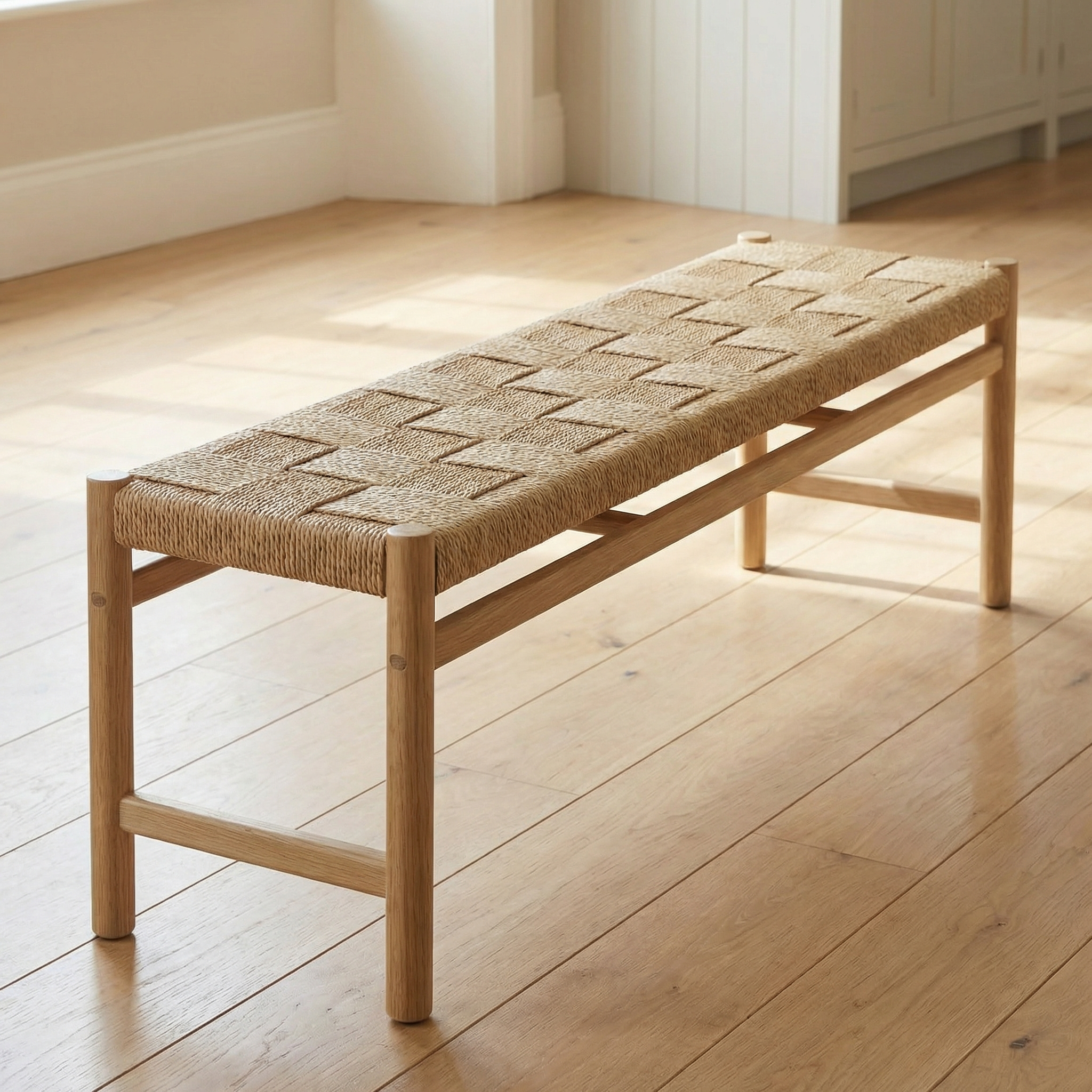The Marlow Woven Dining Bench in Light Oak sits on a light wood floor in a sunlit room.