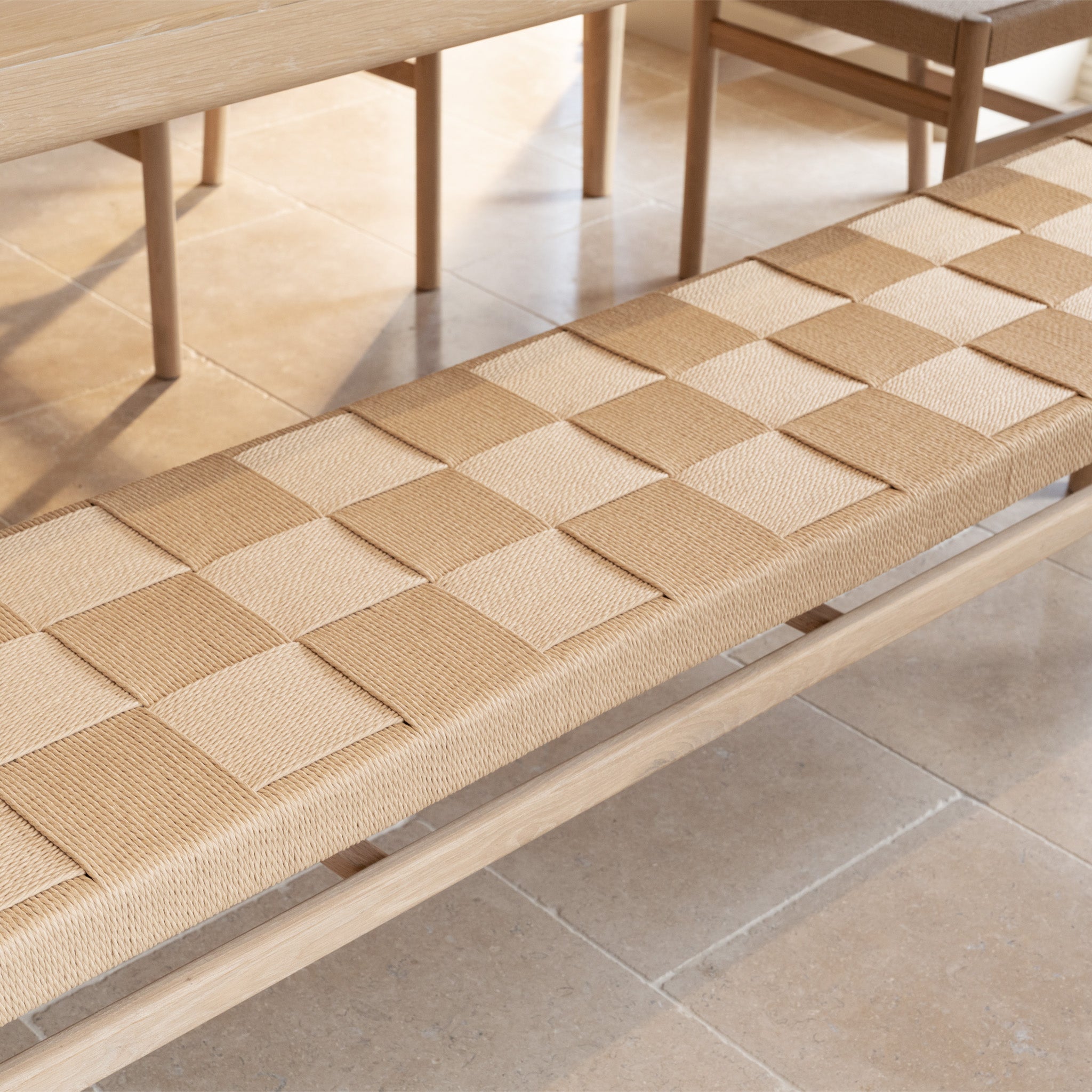 The Marlow Woven Dining Bench in Light Oak, featuring a checkered pattern, sits on a tiled floor next to a wooden table and chairs.