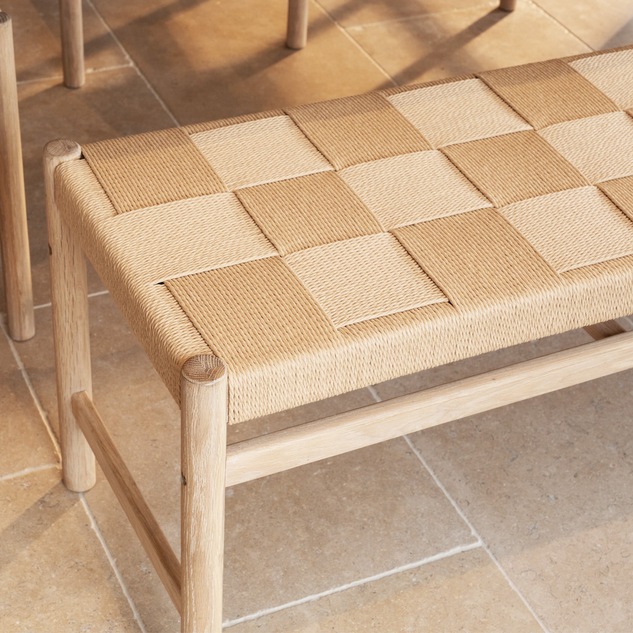 The Marlow Woven Dining Bench in Light Oak, featuring a checkered woven seat, rests on a tiled floor as sunlight casts gentle shadows.