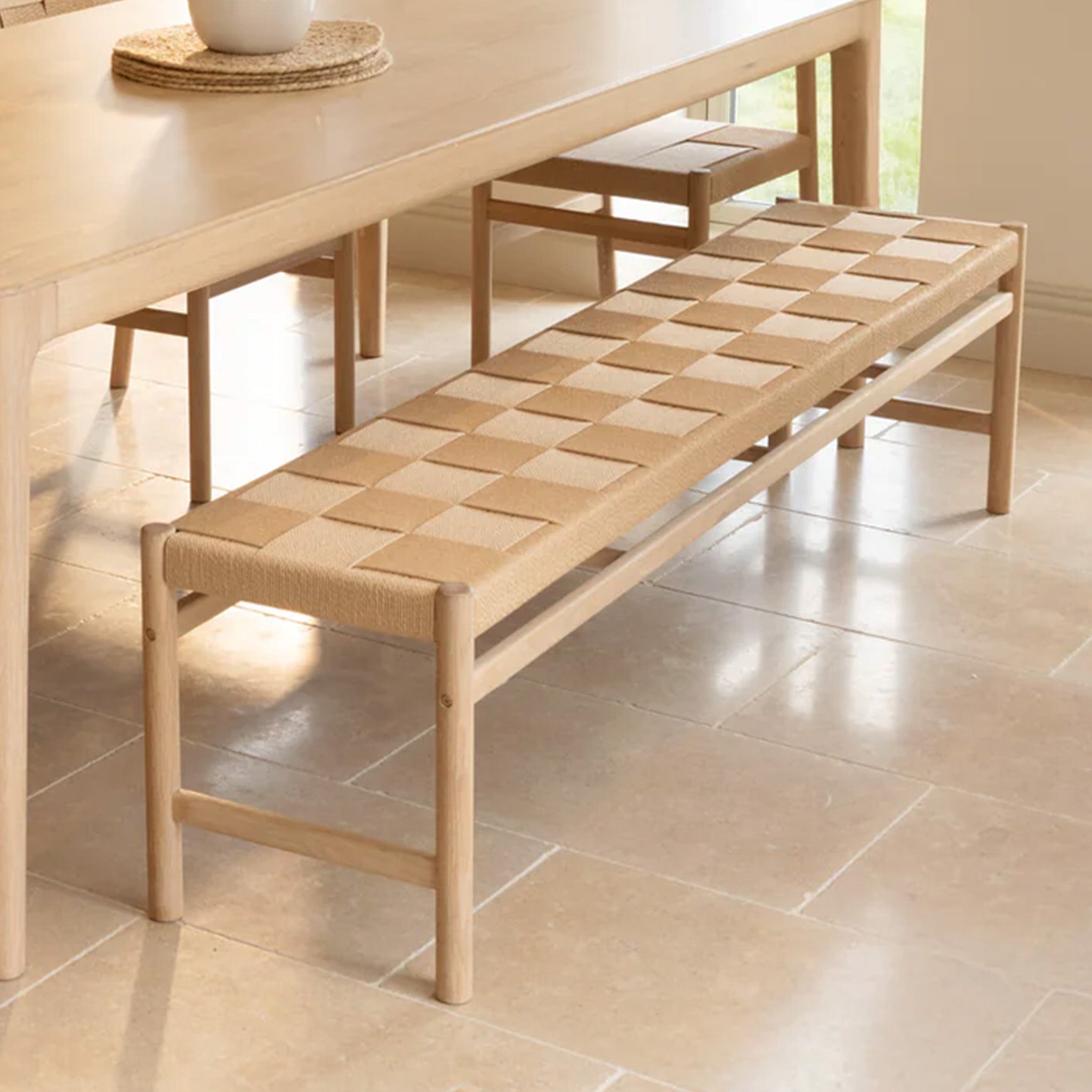 A Marlow Woven Dining Bench in Light Oak sits beside a matching dining table on a tiled floor.