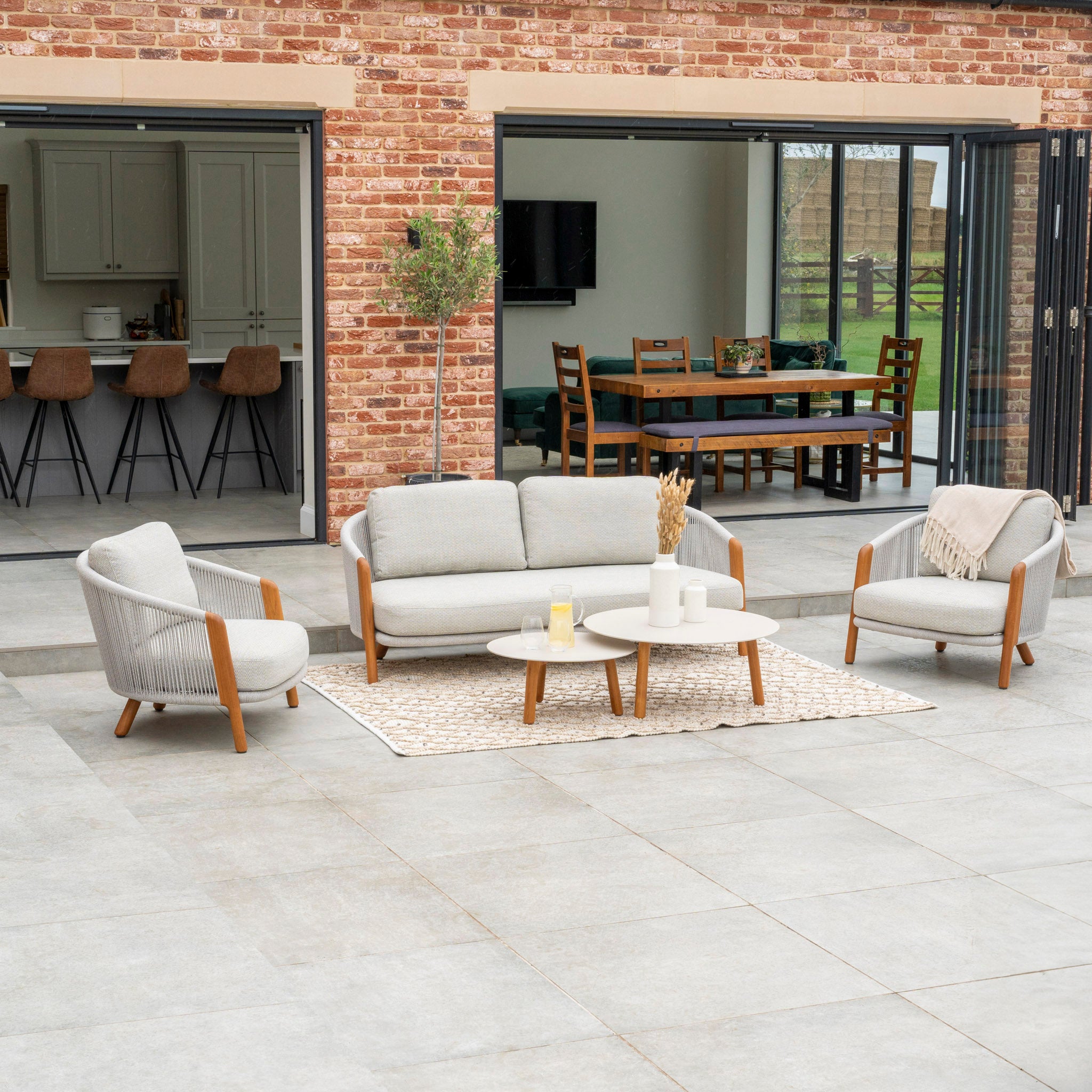 Add a touch of luxury to your outdoor patio area beside your brick house with the Merano 2 Seat Sofa Set and Coffee Tables in Latte. This stylish set includes modern beige chairs, a sofa, a coffee table, and a rug. The hydrophobic olefin rope weave of the Merano collection enhances both elegance and durability for an upscale outdoor experience.