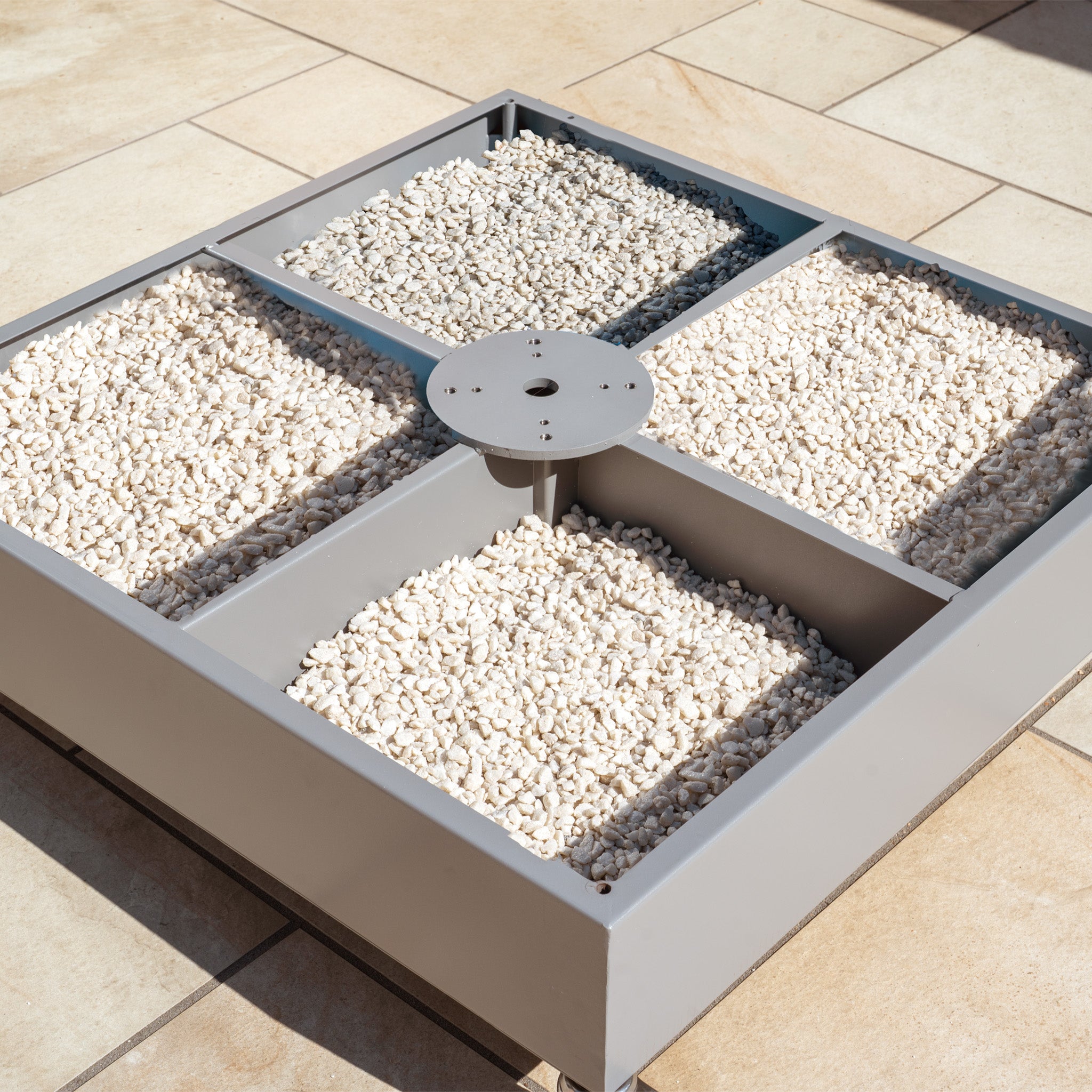 The Fillable Metal Container Parasol Base with Wheels features a stainless steel square base divided into four sections, filled with white stones for stability on tiled floors, making it perfect for securing parasols at outdoor events.