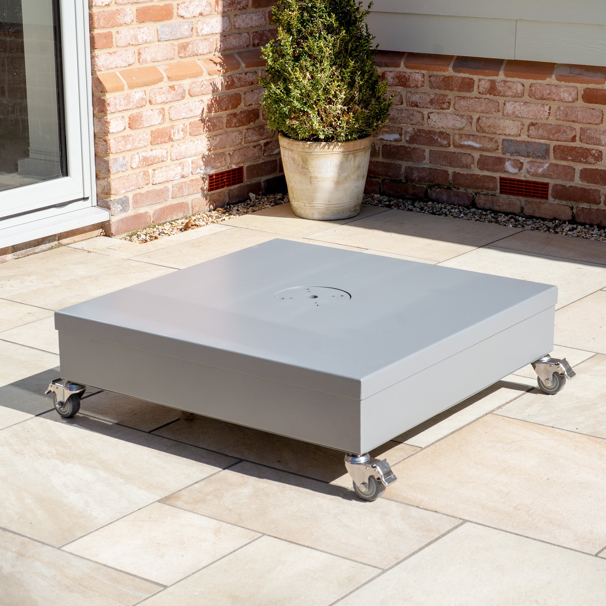 A gray stainless steel Fillable Metal Container Parasol Base with Wheels sits on a patio, complete with a potted plant and brick wall backdrop, ideal for outdoor events.