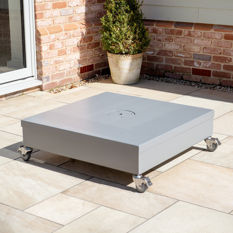 A gray stainless steel Fillable Metal Container Parasol Base with Wheels sits on a patio, complete with a potted plant and brick wall backdrop, ideal for outdoor events.