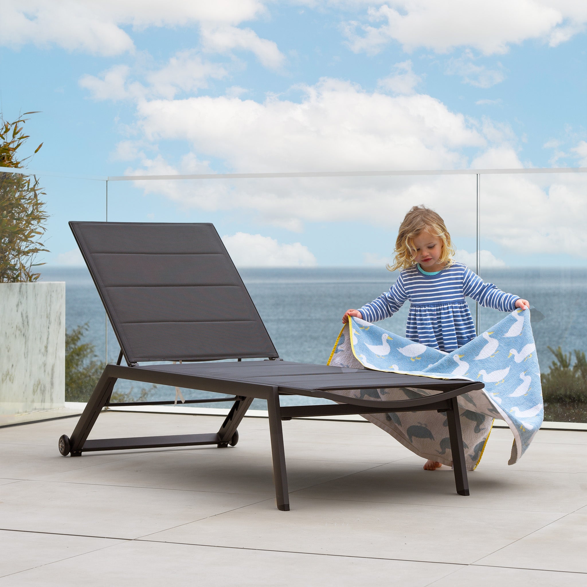 Young child wearing a striped dress holds a blue blanket made of tear-resistant fabric by an outdoor Monaco Padded Sunlounger in Charcoal on a patio overlooking the ocean.
