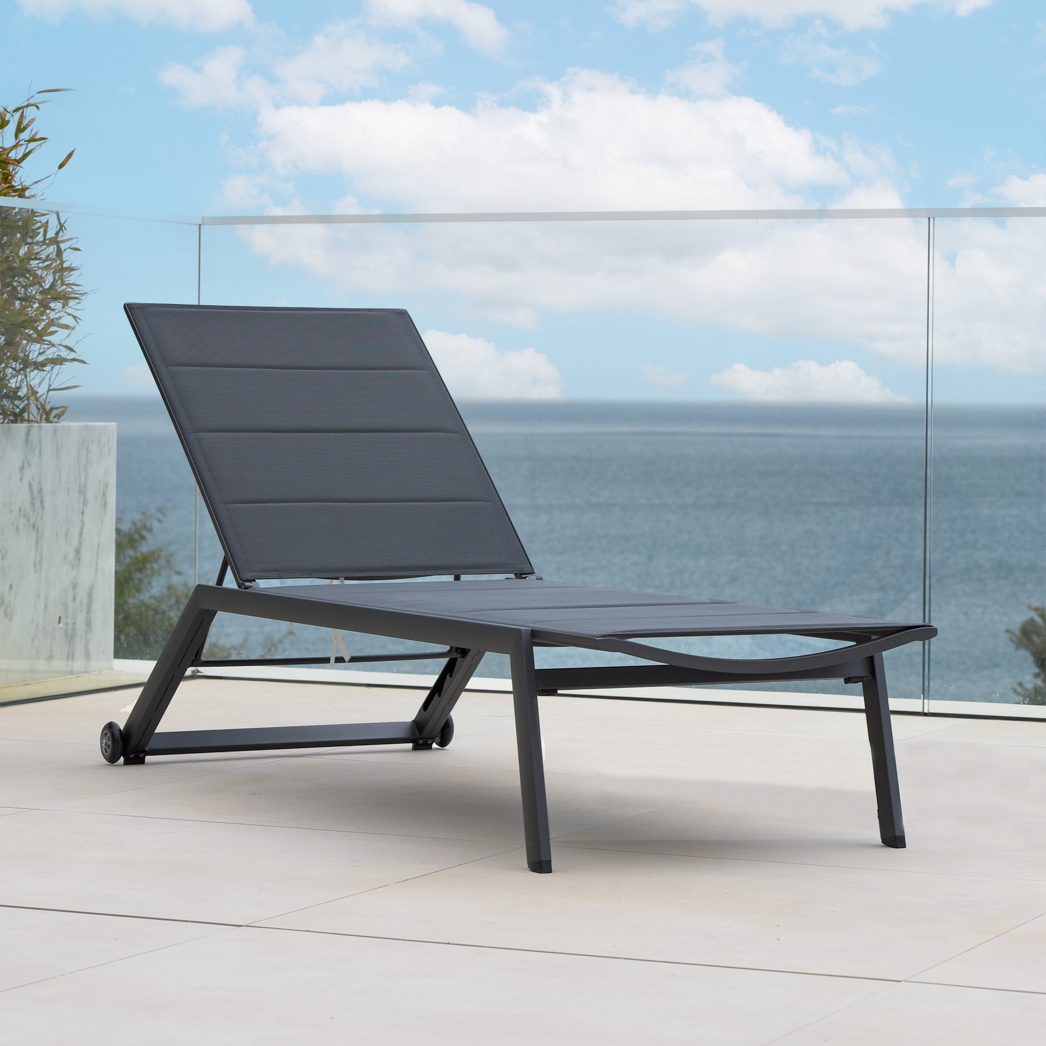 A modern Monaco Padded Sunlounger in Charcoal sits on a patio overlooking the ocean under a partly cloudy sky.