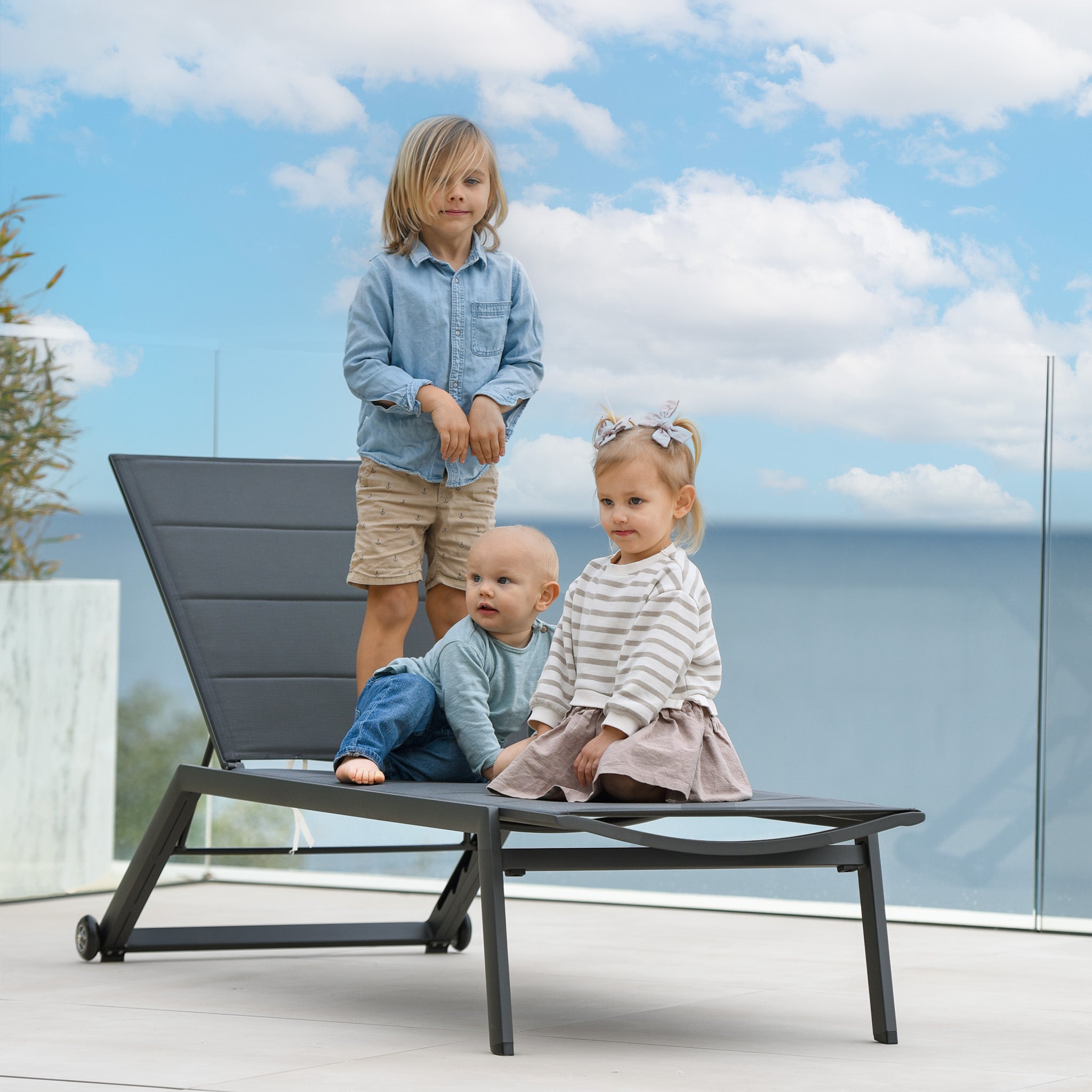 Three children sit and stand on a Monaco Padded Sunlounger in Charcoal outdoors, with the ocean and sky creating a stunning backdrop.