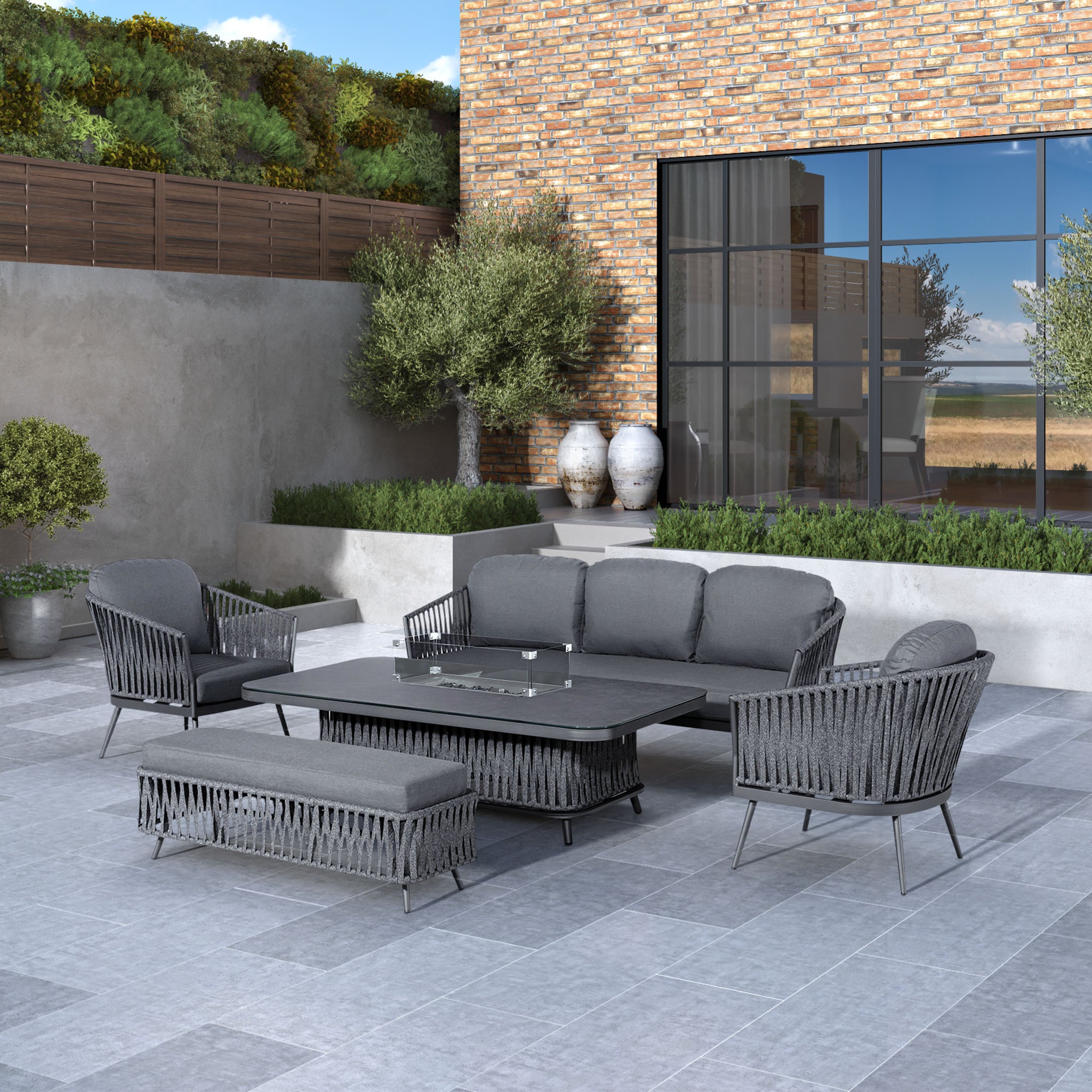 Modern outdoor patio with gray wicker furniture, a Harbour Lifestyle Monterrey rectangular table, and benches set against a brick and glass building. A cozy Monterrey 3 Seat Rope Sofa Set with Rising Firepit Table in Grey surrounds the built-in firepit for an inviting atmosphere.