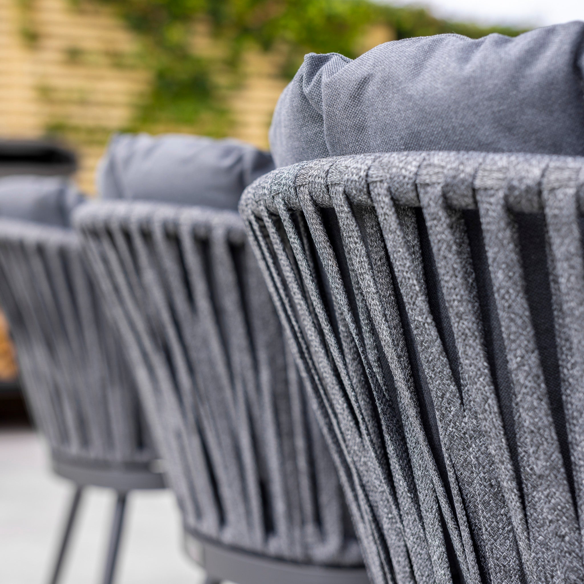 Close-up of Monterrey 8 Seat Rope Rectangular Ceramic Firepit Dining Set in Grey, showcasing grey woven chairs with cushions, lined up in a row. The chairs feature high-quality aluminium construction for durability and style, with greenery in the background.