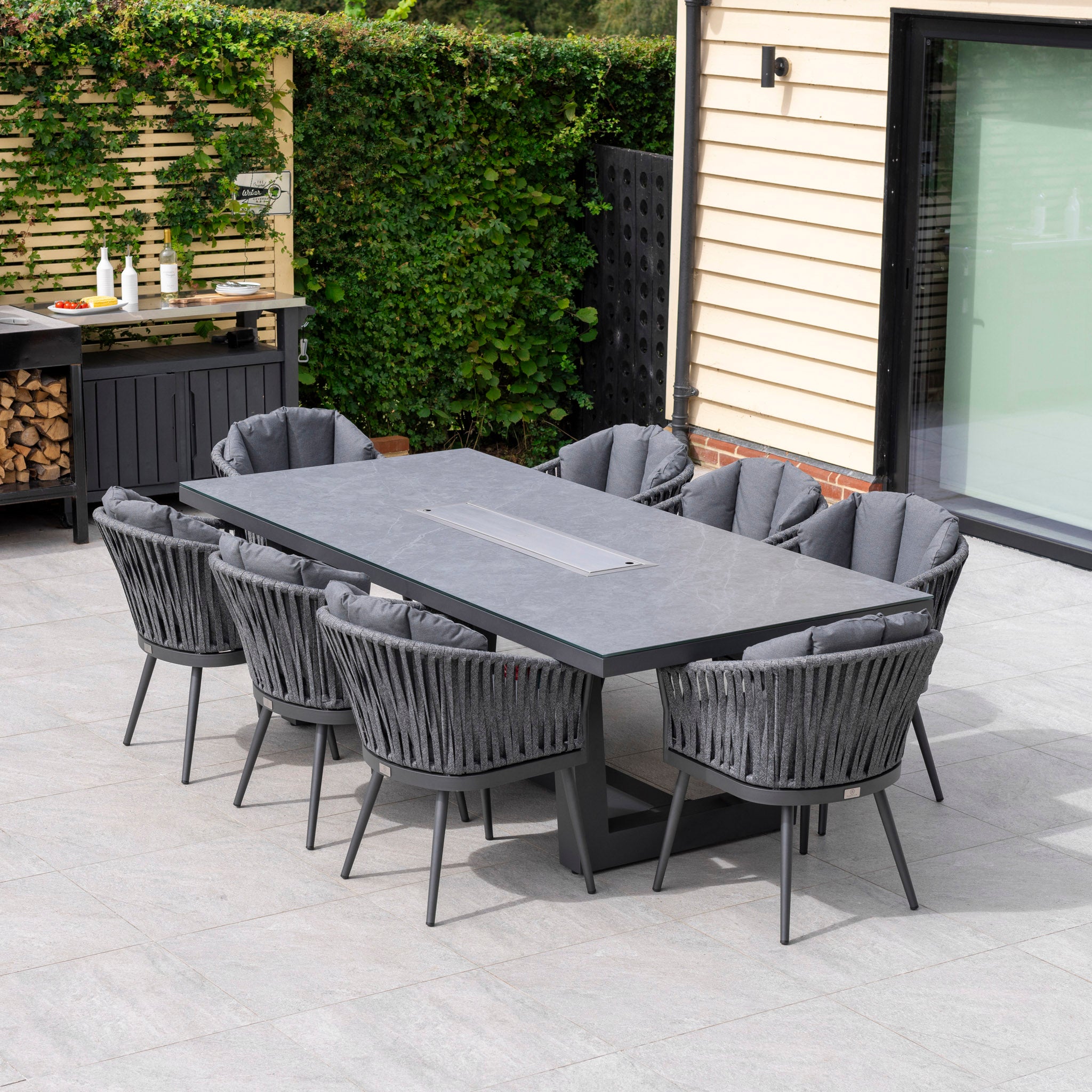 Outdoor patio with a Harbour Lifestyle Monterrey 8 Seat Rope Rectangular Ceramic Firepit Dining Set in Grey, including eight padded chairs. The high-quality aluminum rectangular table and barbecue grill are near a fence adorned with lush greenery.