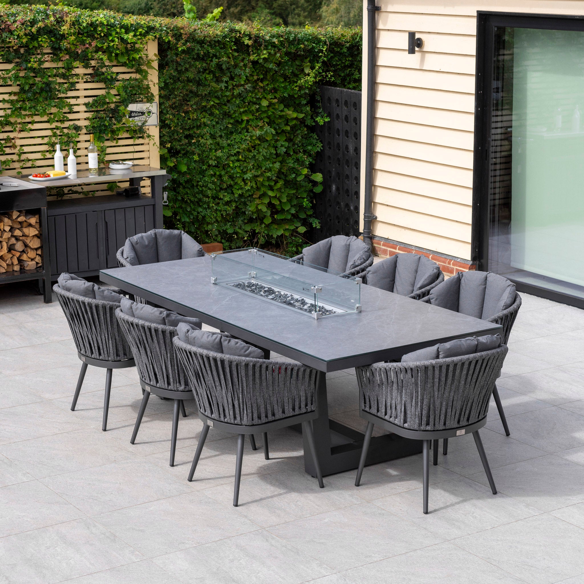 The Monterrey 8 Seat Rope Rectangular Ceramic Firepit Dining Set in Grey from Harbour Lifestyle features a high-quality aluminum table and eight cushioned grey chairs, complete with a central glass-protected fire pit. This stunning dining set is beautifully accentuated by lush greenery in the background.