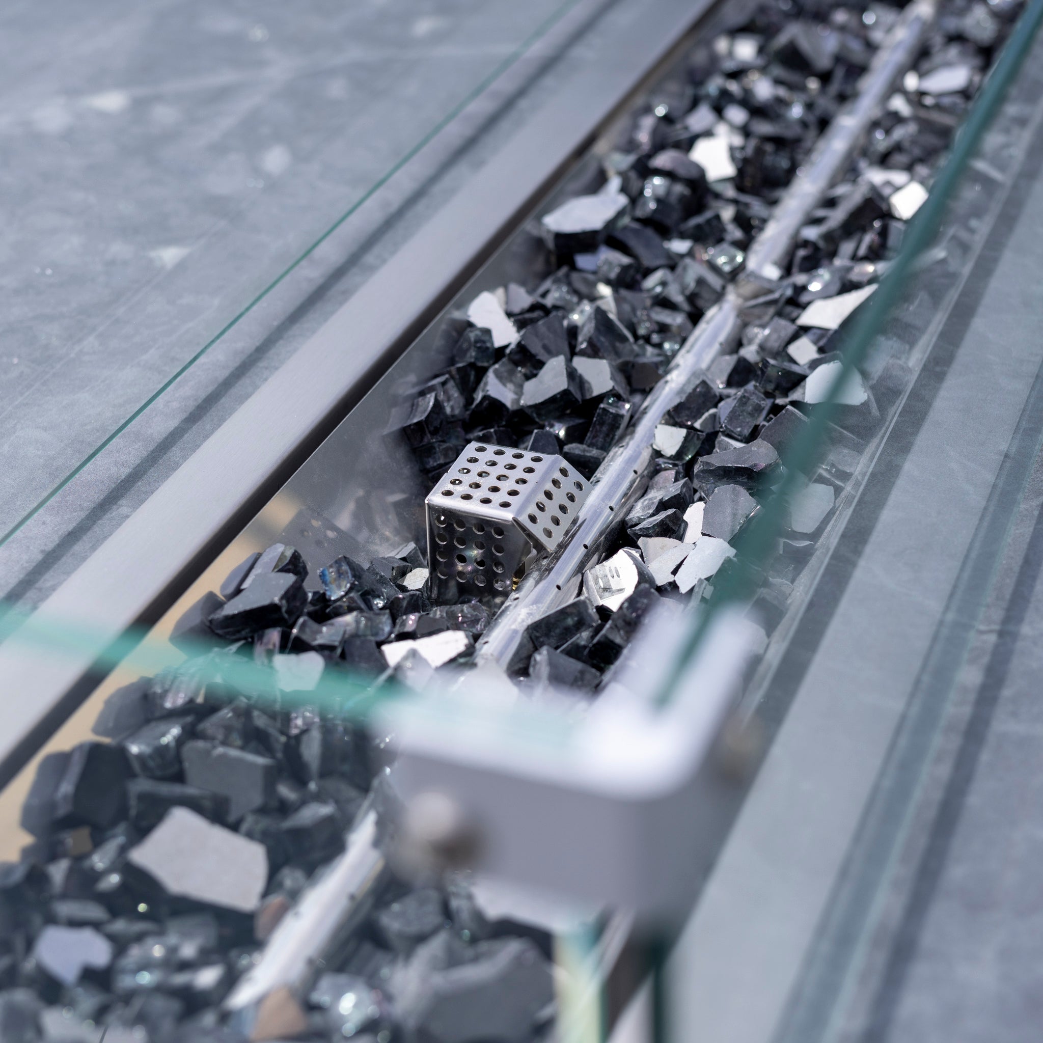A metal die amid shattered black rocks in a narrow glass container, reminiscent of the sophisticated design found in the Monterrey 8 Seat Rope Rectangular Ceramic Firepit Dining Set in Grey, crafted from high-quality aluminium.