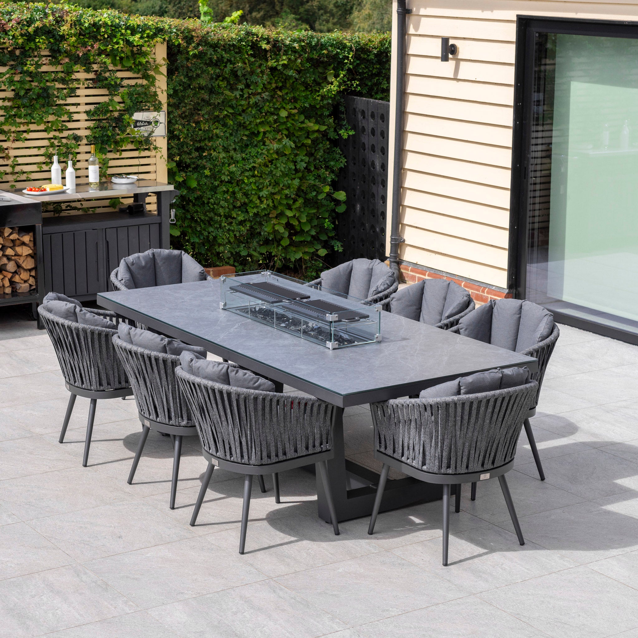 Modern outdoor dining area with a long table and eight cushioned chairs on a patio, surrounded by greenery. The Monterrey 8 Seat Rope Rectangular Ceramic Firepit Dining Set in Grey adds elegance and functionality to your outdoor space.