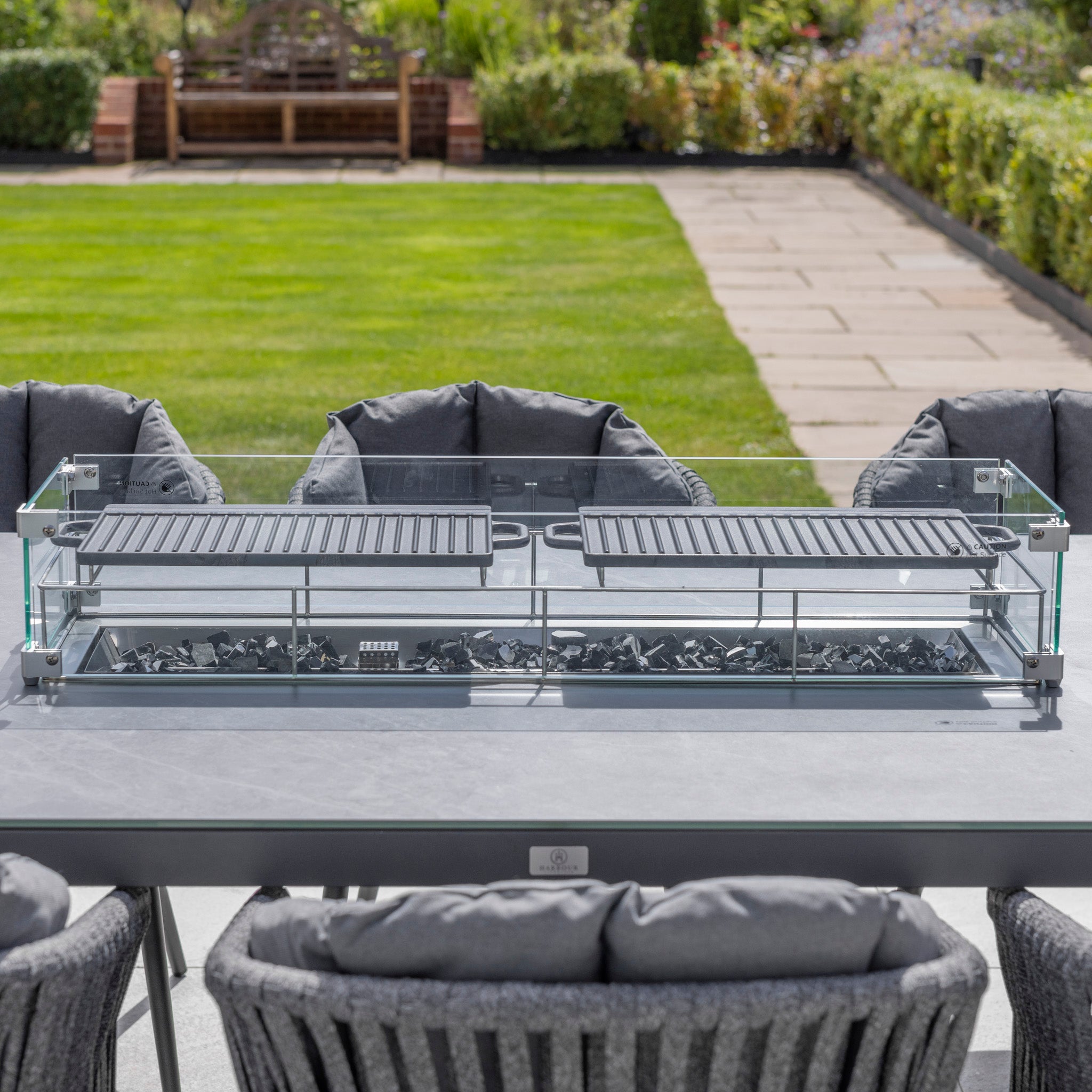 The Monterrey 8 Seat Rope Rectangular Ceramic Firepit Dining Set in Grey from Harbour Lifestyle features a long table with a glass-enclosed grill and grey cushioned chairs, all crafted from high-quality aluminium, perfect for an elegant garden setting.