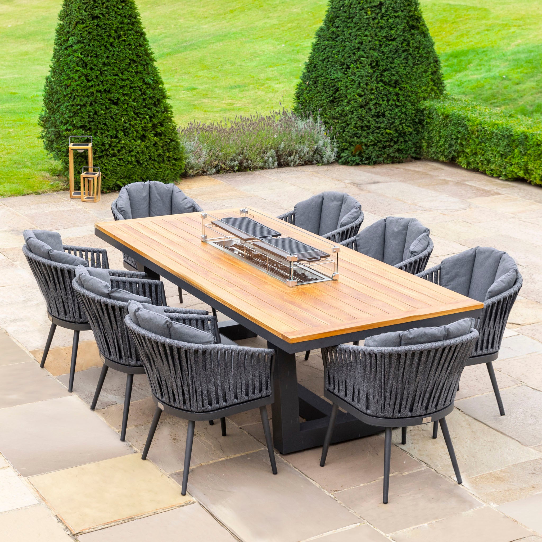 Monterrey 8 Seat Rope Rectangular Teak Firepit Dining Set in Grey on a stone patio, surrounded by greenery.