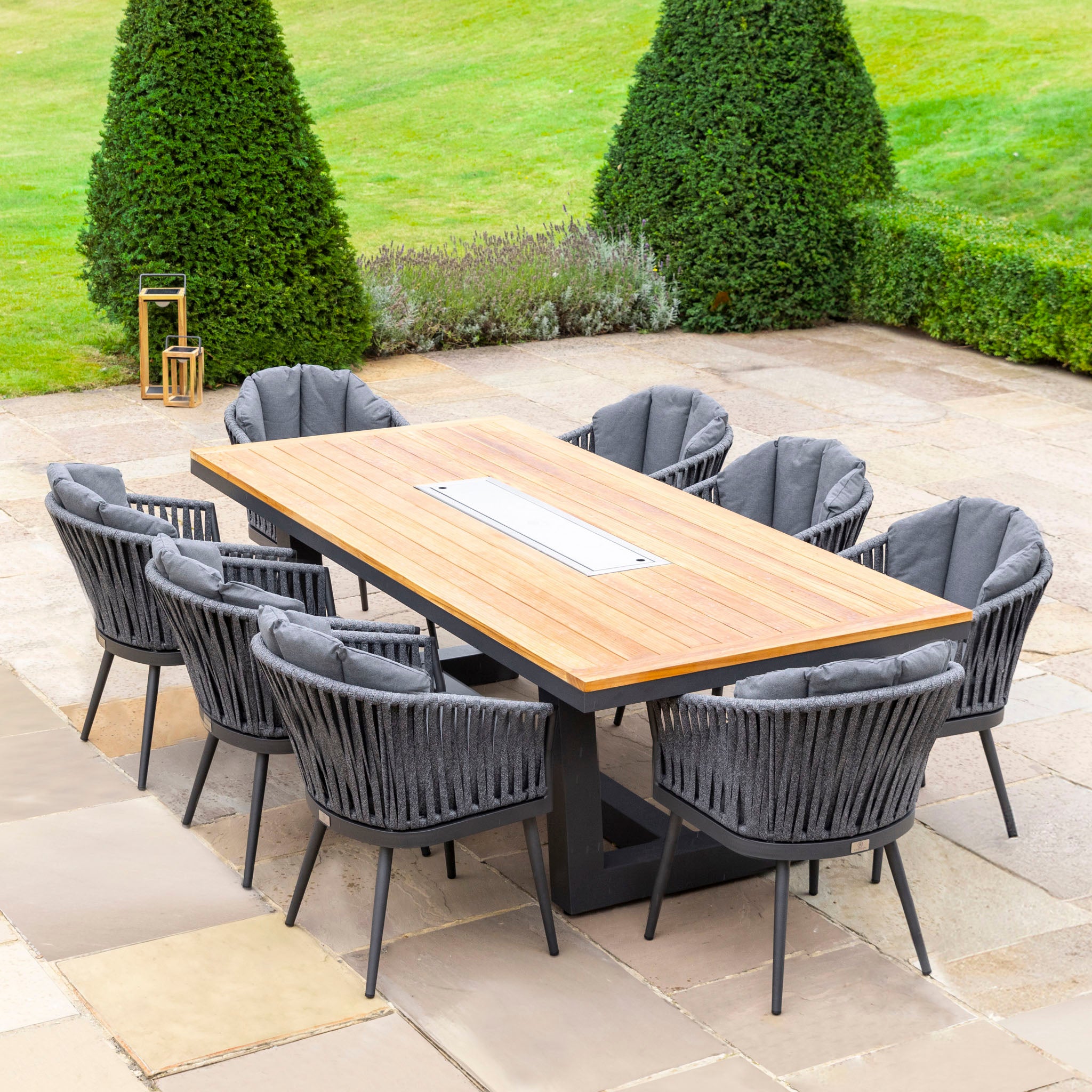 Experience luxury with the Harbour Lifestyle Monterrey 8 Seat Rope Rectangular Teak Firepit Dining Set in Grey, featuring a sleek wooden table and eight plush gray cushioned chairs, all perfectly complemented by lush greenery. For an elevated ambiance on cooler evenings, enjoy the built-in firepit as an added feature.