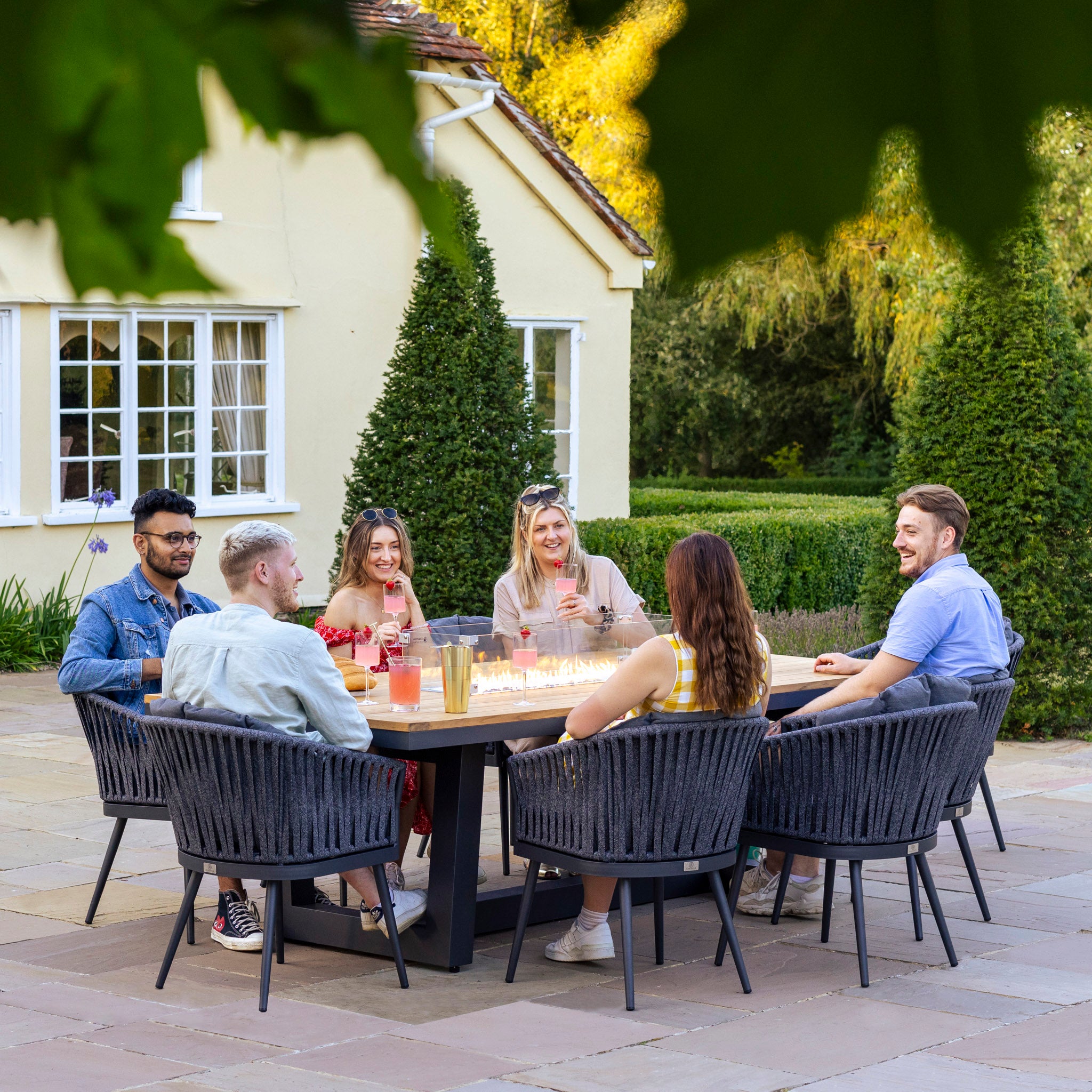 Six people are sitting around a Monterrey 8 Seat Rope Rectangular Teak Firepit Dining Set in Grey, enjoying drinks and chatting near a house with trees in the background.