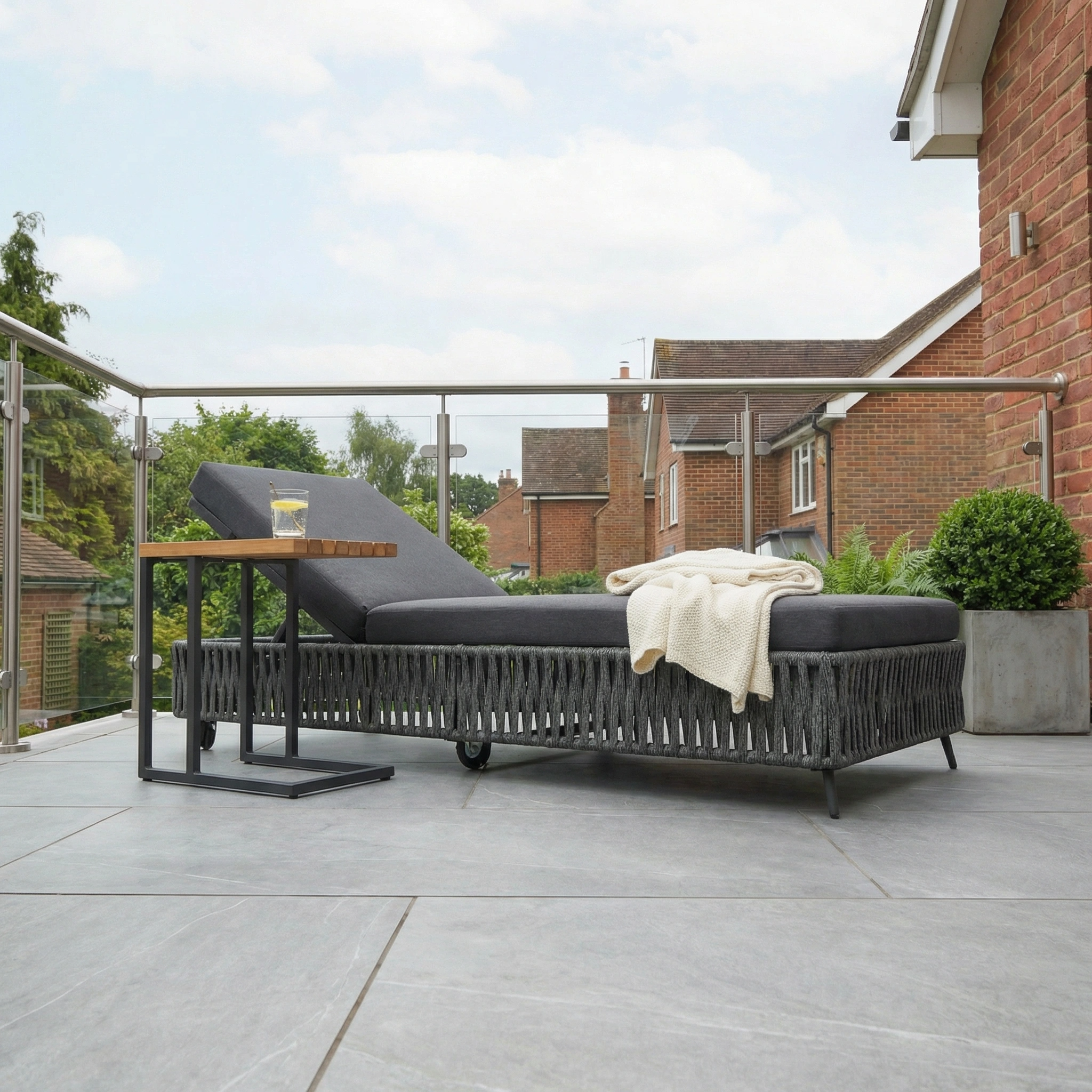 The Monterrey Rope Sun Lounger in Grey sits on a tiled patio beside a brick house, paired with a blanket, side table, and drink.