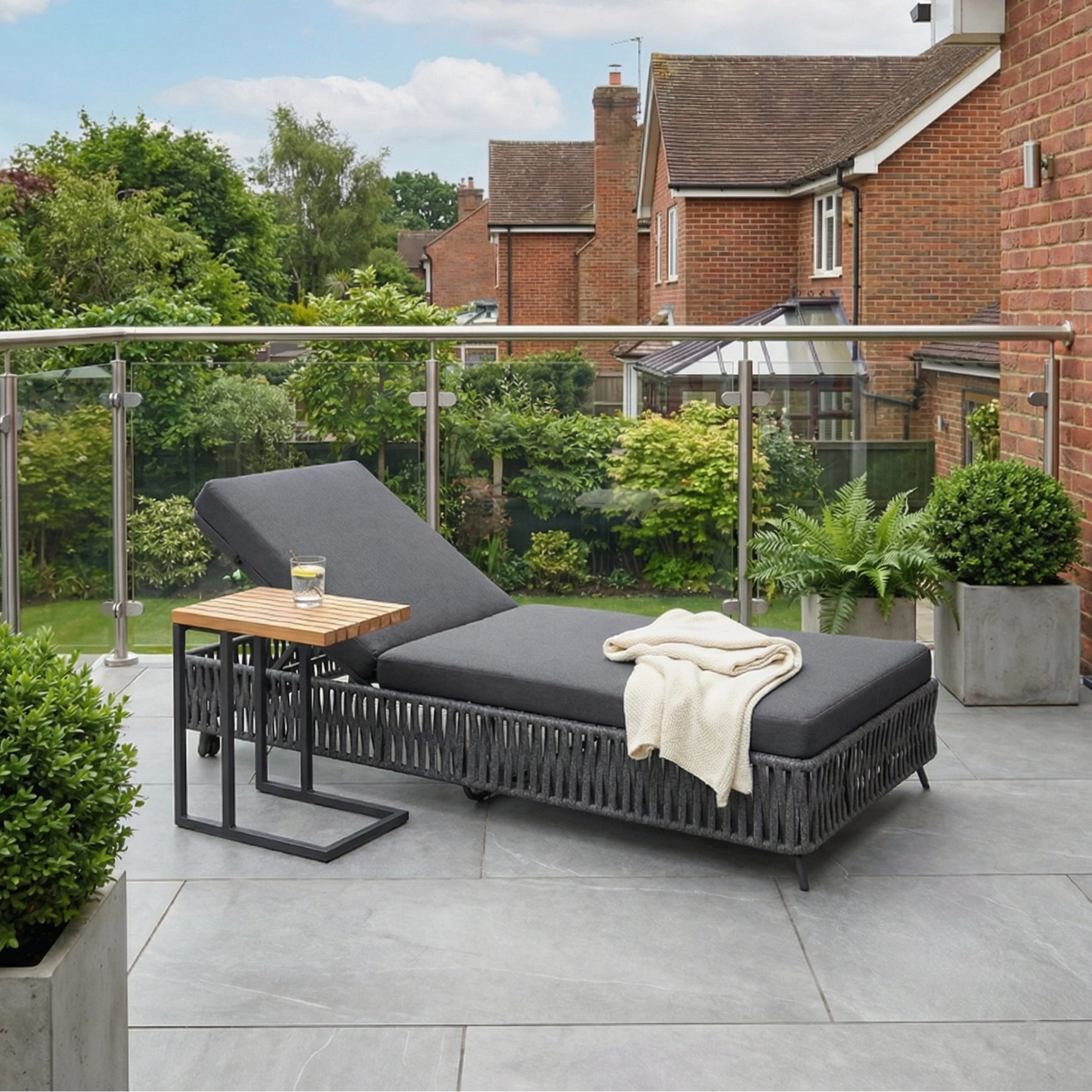 Monterrey Rope Sun Lounger in Grey with weatherproof cushions and a white blanket on a patio, paired with a side table and potted plants—ideal outdoor furniture for relaxing in style.