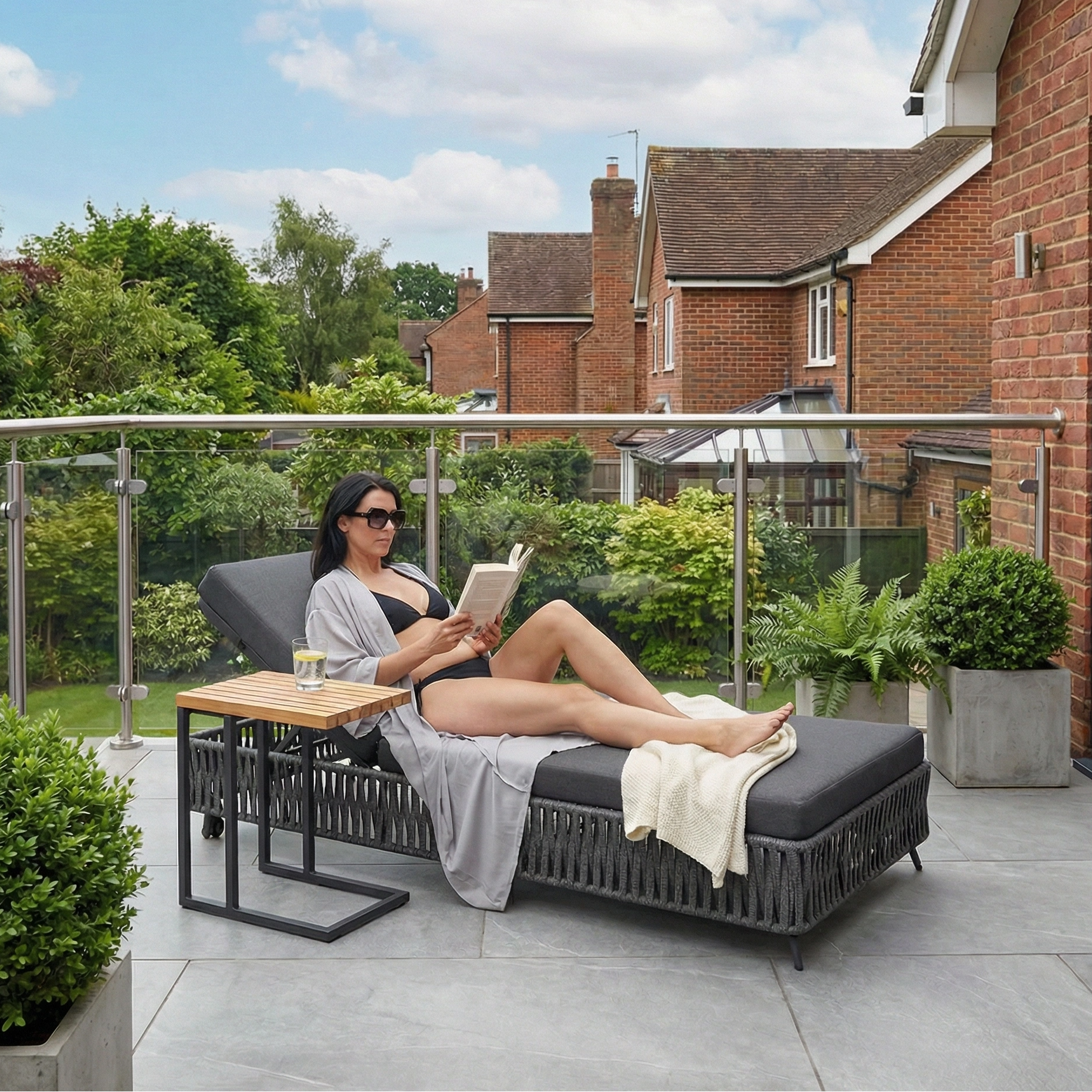 A woman relaxes on the Monterrey Rope Sun Lounger in Grey, reading a book on the patio with a drink at her side, surrounded by greenery and enjoying the comfort of its weatherproof cushions.