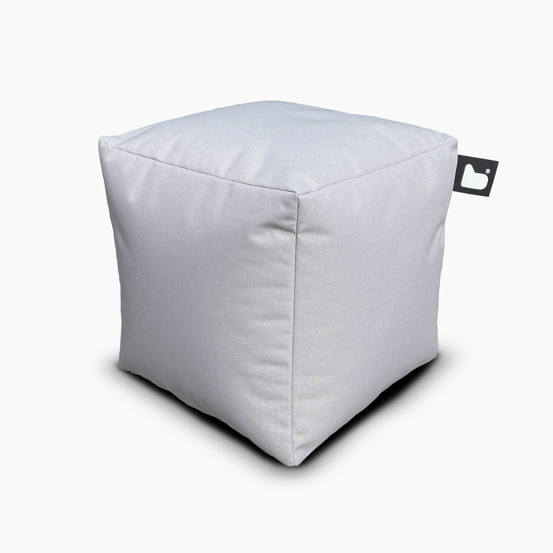 The Outdoor B-Box in Silver Grey is a cube-shaped fabric pouf designed for extreme lounging, featuring a small black tag on one corner displaying a white heart icon. Perfect for both indoor and outdoor spaces, this versatile piece embodies style and comfort.