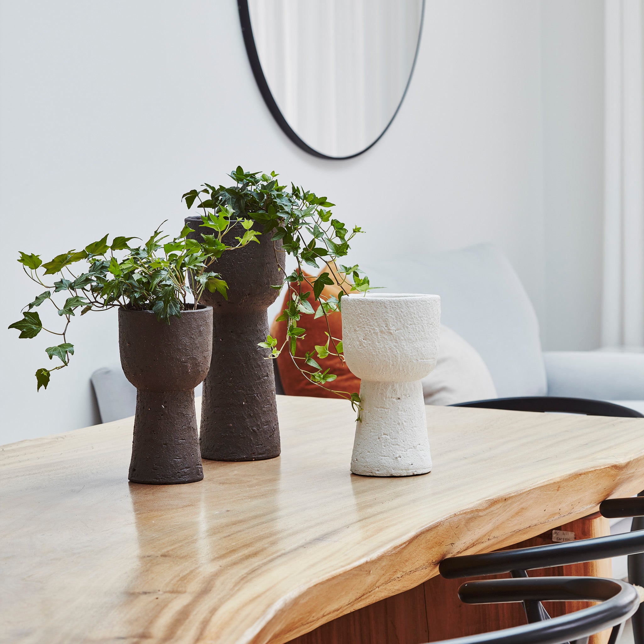 Three textured vases with green vines sit on a wooden table, accompanied by the rustic mocha exterior of an Ivyline Nico Tall Planter. In the background, a round mirror reflects the serene setting alongside a cozy sofa, blending beautifully into this European-inspired scene.