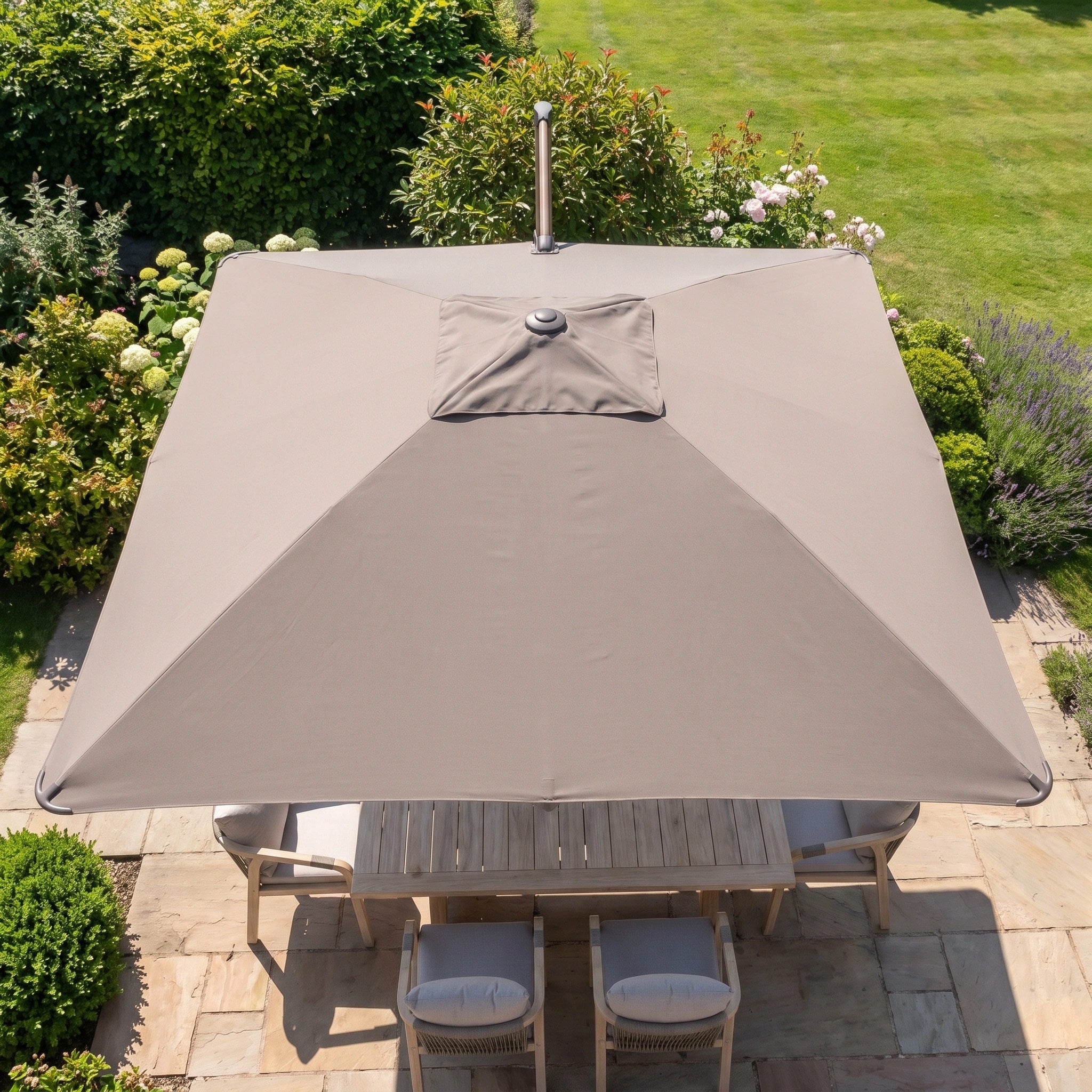The Nexus T2 Premium 3m Square Cantilever Parasol in Deep Taupe, supported by the Venezia 90kg Granite Base, provides shade for a wooden table and contemporary garden furniture on a stone patio, all set within a lush green garden.