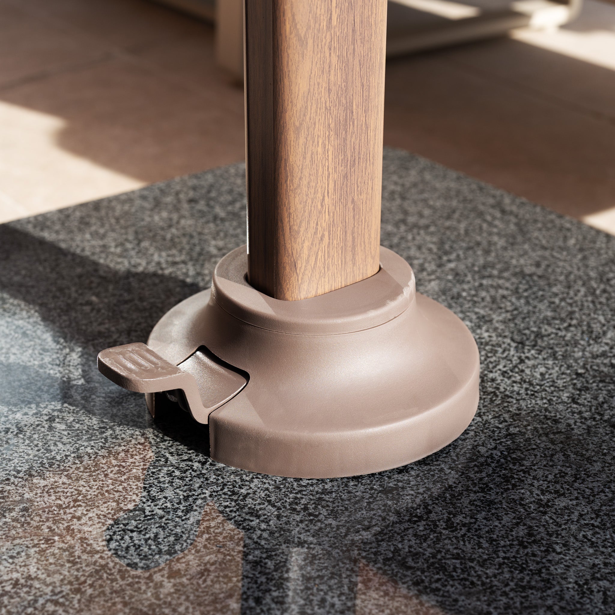 The Nexus T2 Premium 3m Square Cantilever Parasol in Deep Taupe with a 90kg granite base rests on speckled gray stone tiles in bright sunlight—ideal for enhancing your garden furniture setup.