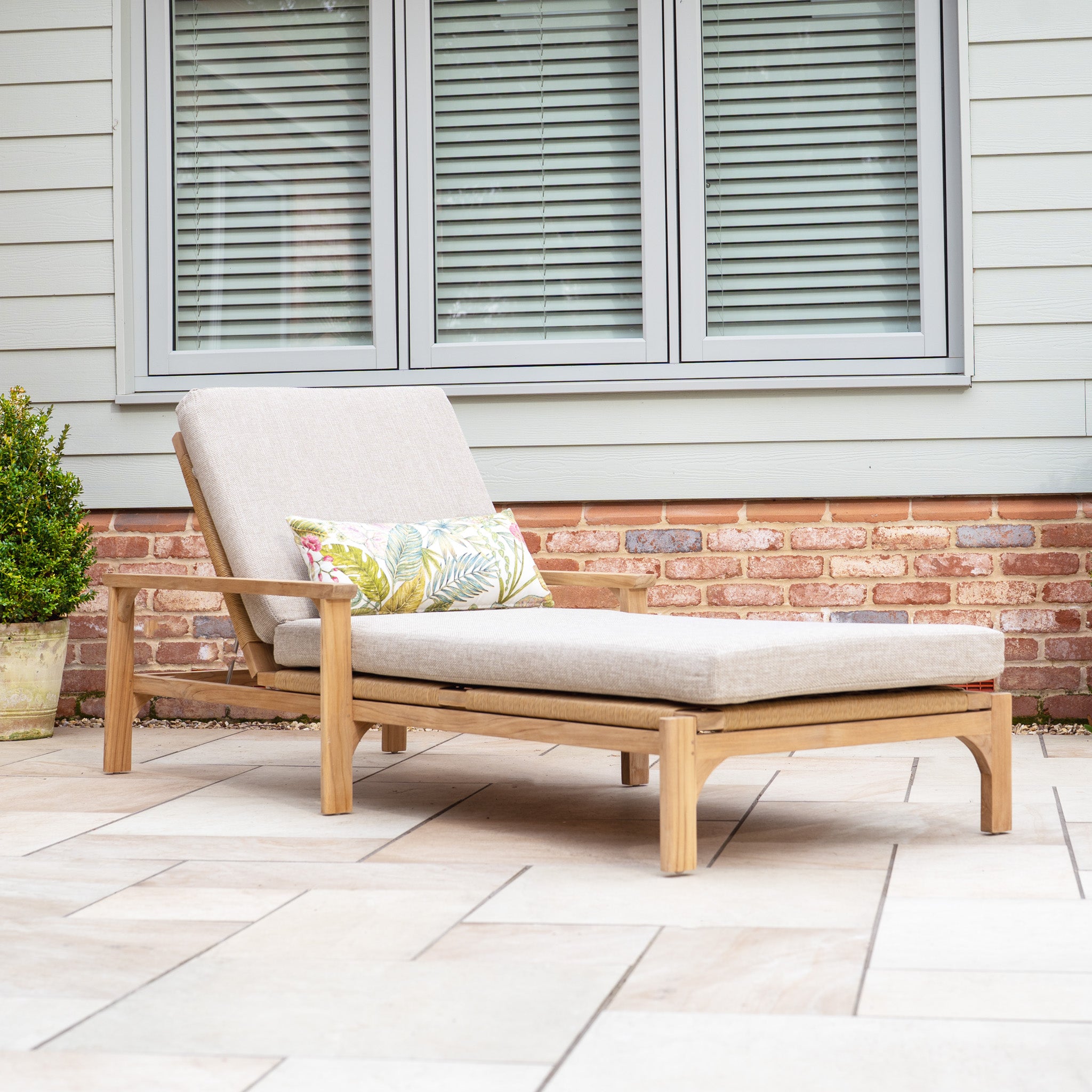 The Nola Sunlounger in Fawn, made from premium teak wood, includes a plush cushion and floral pillow, ideal for enhancing any patio space elegantly positioned by a window with closed blinds.
