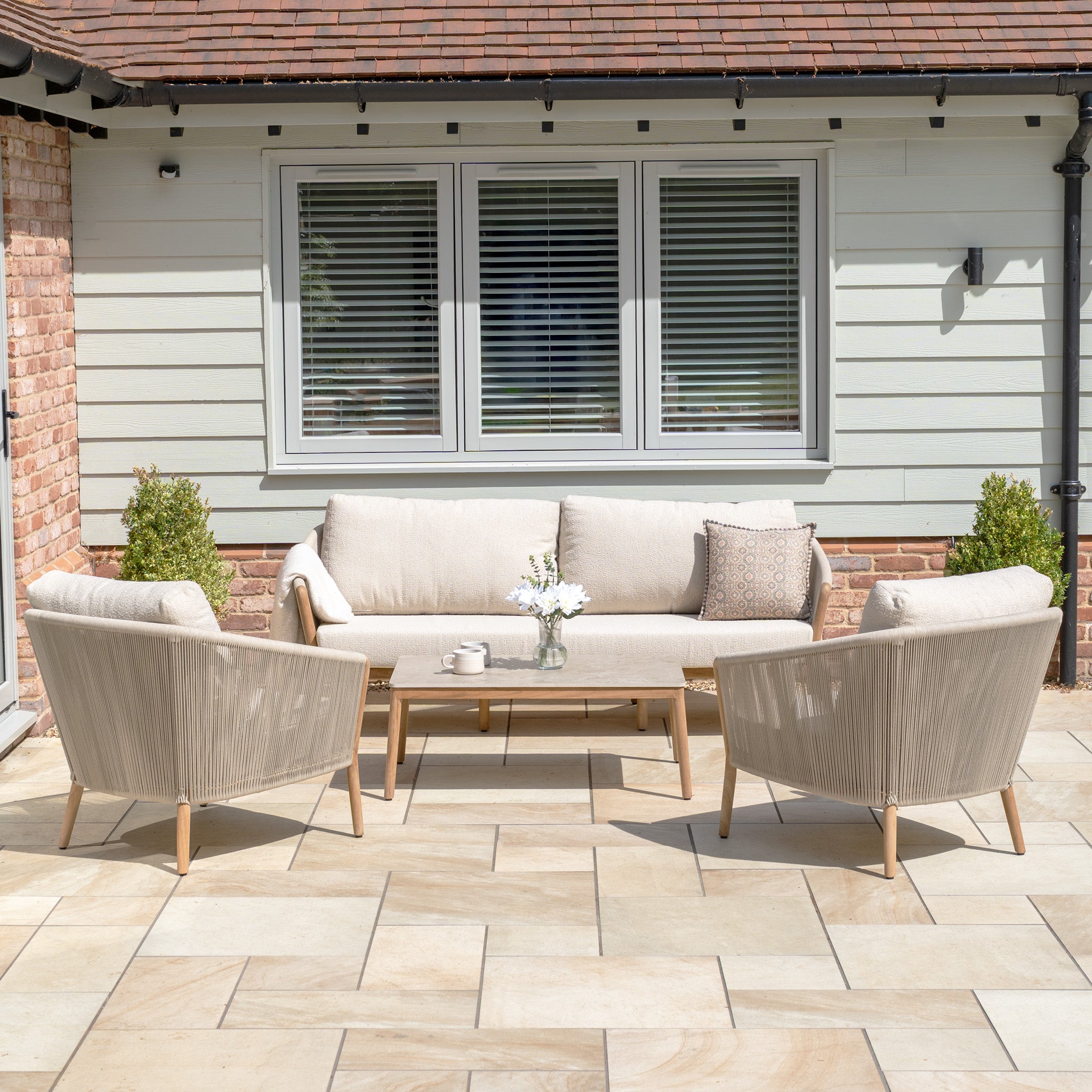 The New York 3 Seat Sofa Set includes two chairs and a table with flowers in a beige, weather-resistant design—ideal for adding style to your stone-tiled patio.