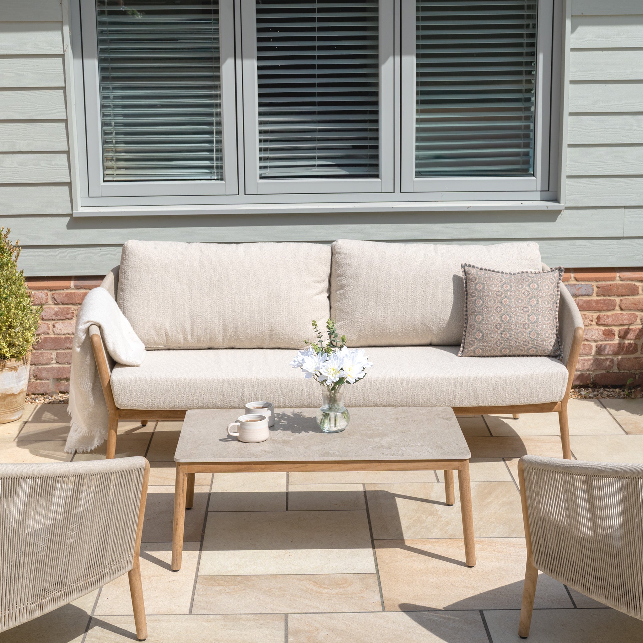 The New York 3 Seat Sofa Set, shown with a coffee table, mug, and flowers on a patio by a window, offers stylish, weather-resistant outdoor furniture—ideal for gatherings.