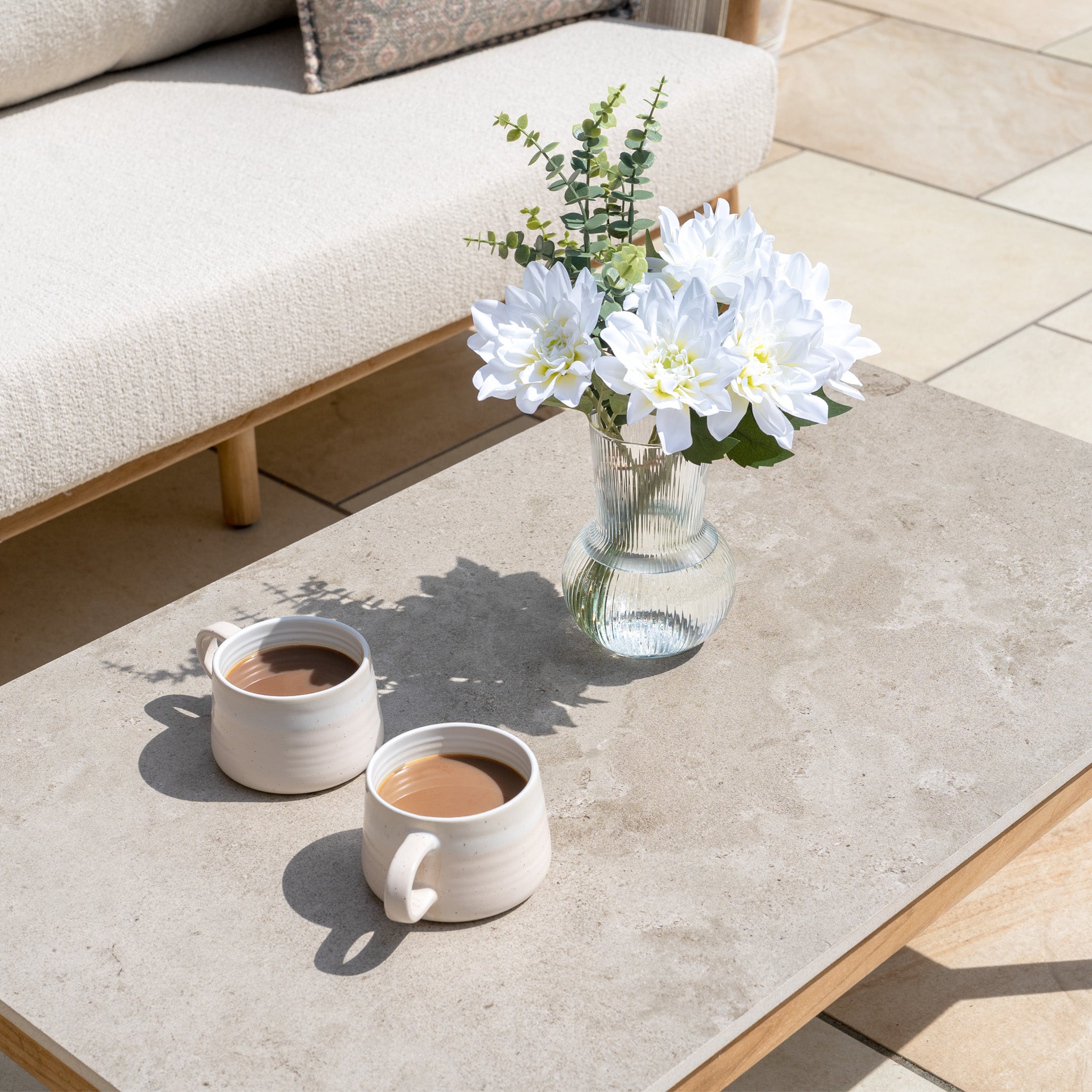 Two mugs of coffee and a white flower vase sit on a light stone table next to the weather-resistant New York 3 Seat Sofa Set, making a cozy outdoor retreat.