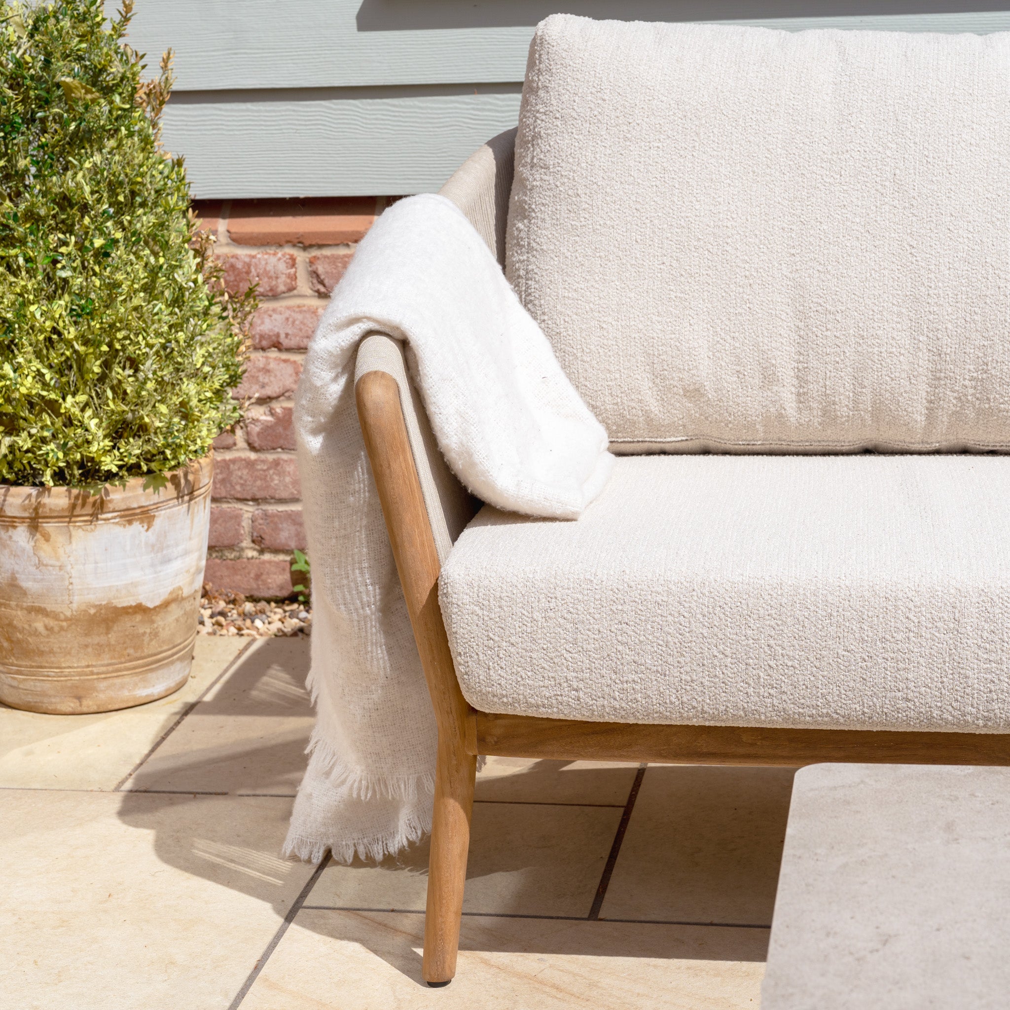 The New York 3 Seat Sofa Set, shown with a white throw on the arm next to a potted plant on a sunny patio, offers weather-resistant comfort and style for relaxing outdoors.