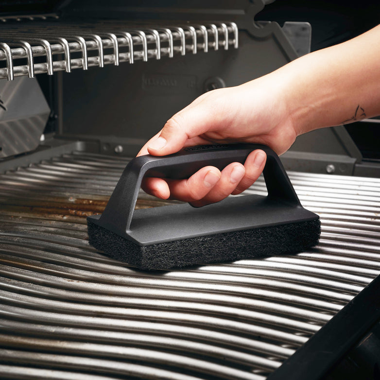 A hand holding a Napoleon Abrasive BBQ Scrubber is cleaning the porcelainized cast iron cooking grids of a grill.