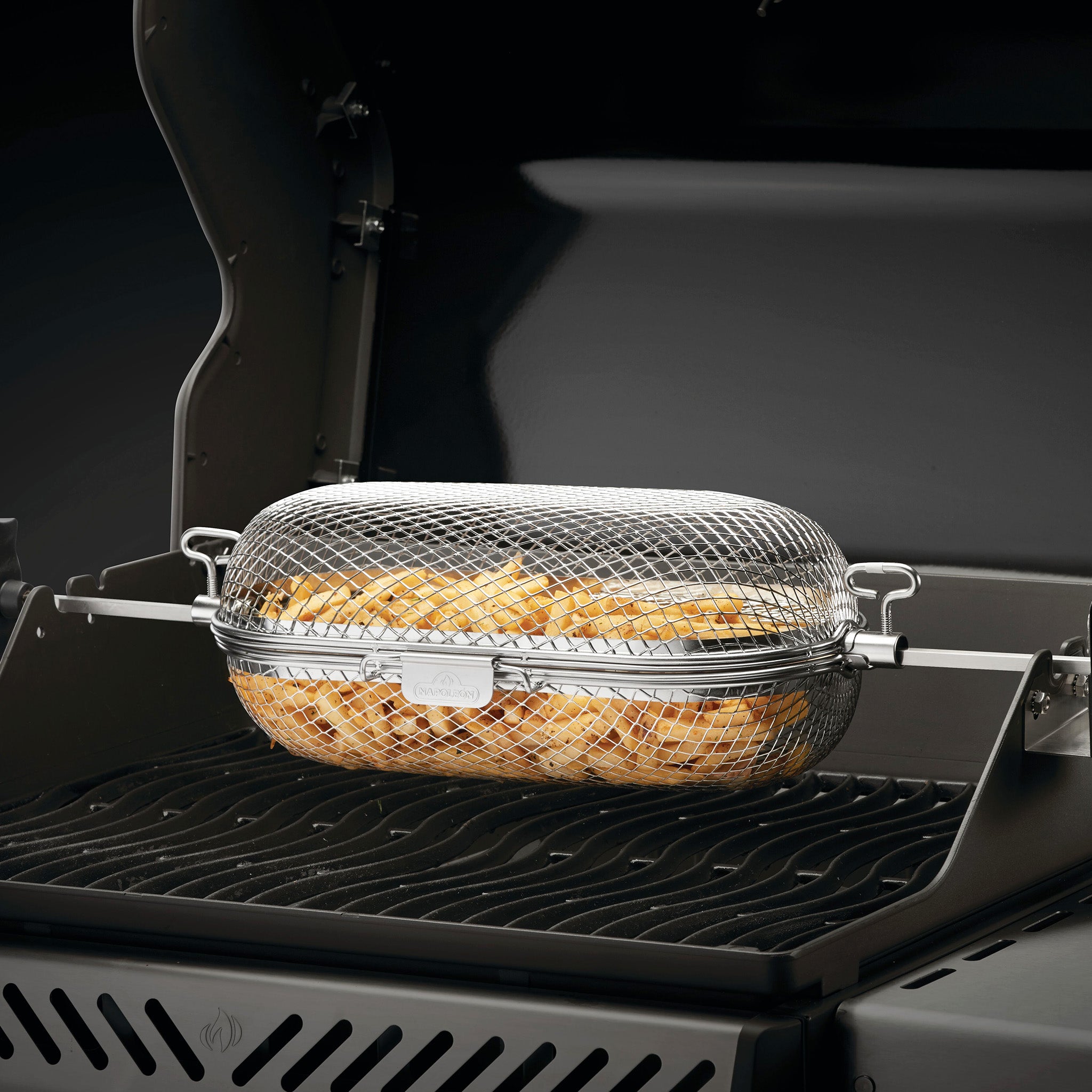 A Napoleon Stainless Steel Rotisserie Basket filled with sliced potatoes is placed on a barbecue grill with the lid open.