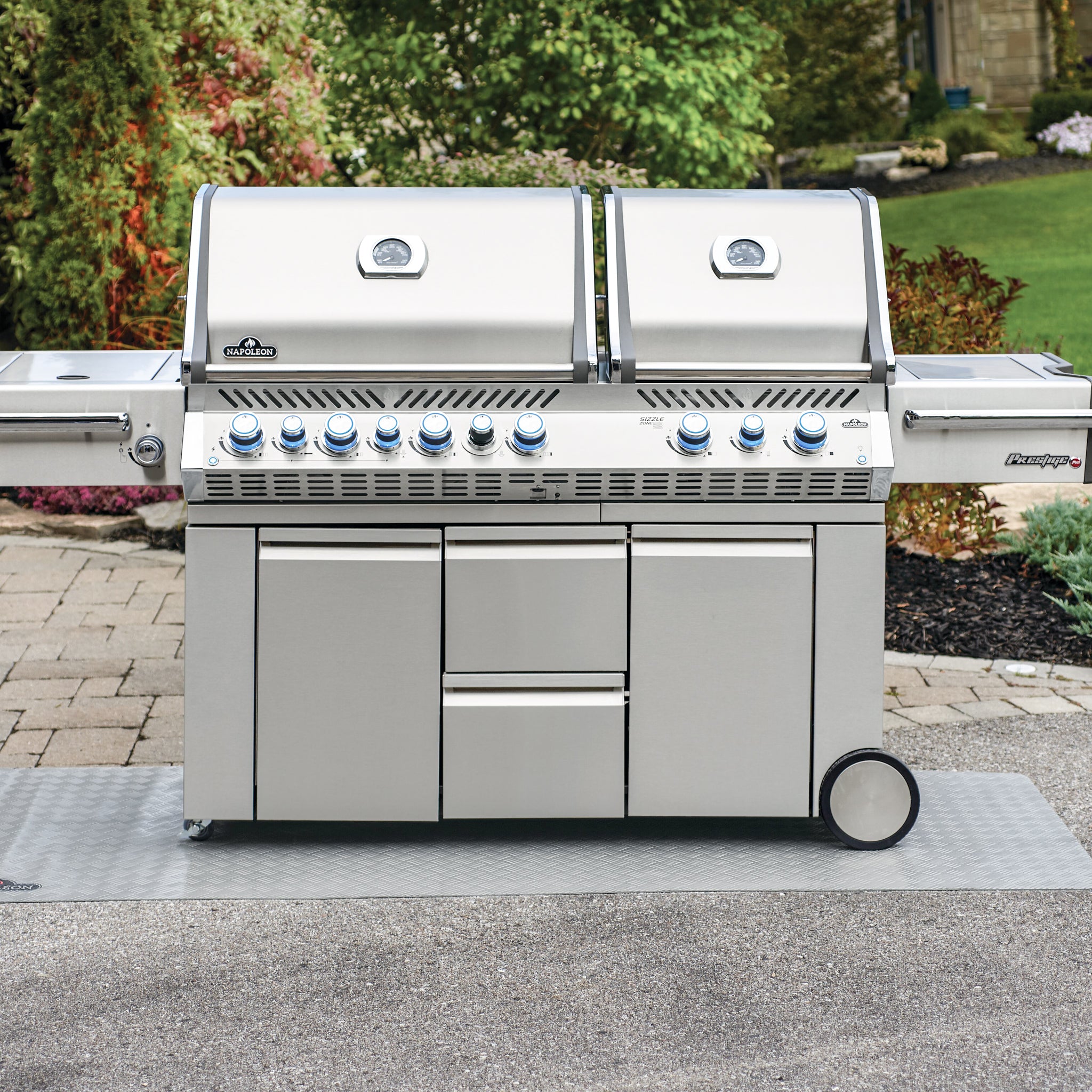 A large stainless steel outdoor grill with multiple control knobs, dual lids, and storage cabinets stands on a paved area, protected underneath by a Napoleon Grill Mat - Large.