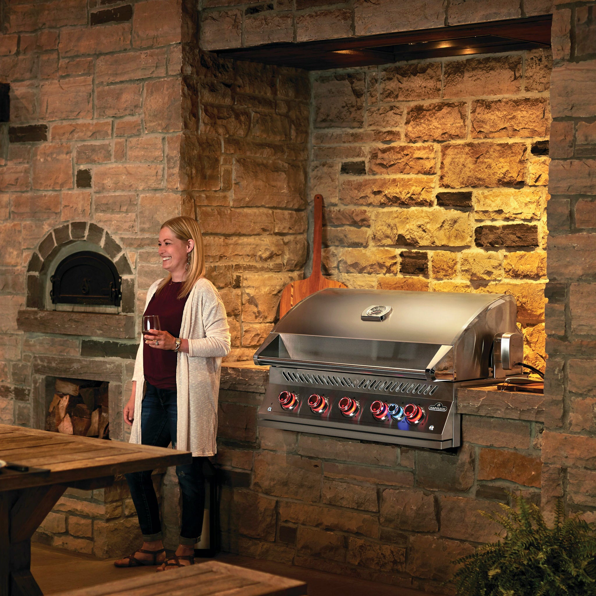 A woman holding a glass stands near an outdoor stone brick kitchen, admiring the Napoleon 700 Series 38" Built-in Gas Grill equipped with a high-performance JETFIRE ignition system and rear infrared burner.
