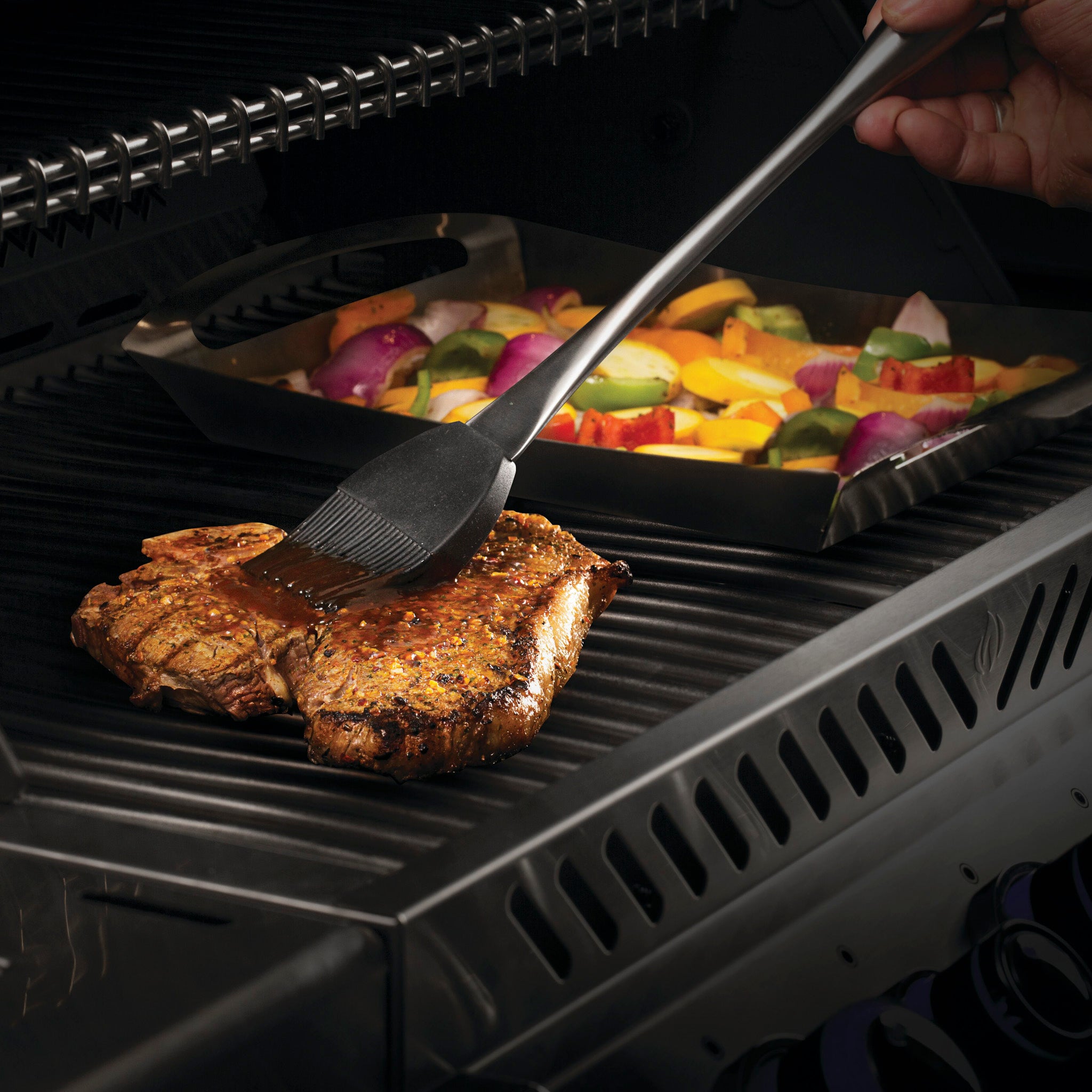 A person bastes a steak on a grill with a tray of assorted vegetables nearby, using the Napoleon 3-Piece PRO Stainless Steel Grill Toolset for precision and ease.
