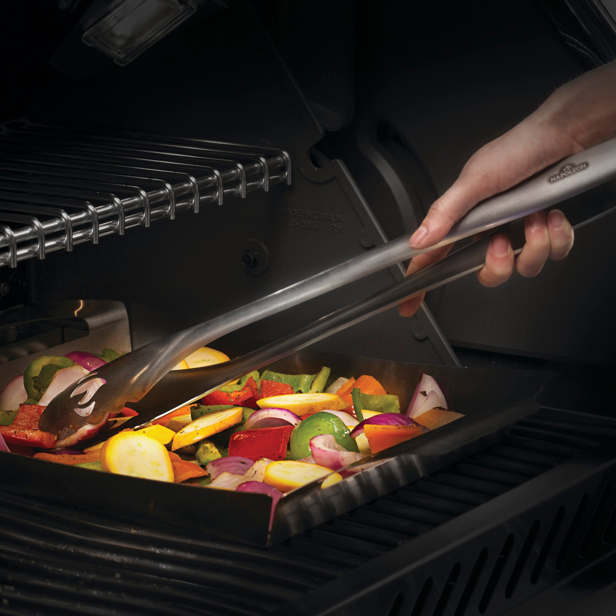 A hand holding tongs from the Napoleon 3-Piece PRO Stainless Steel Grill Toolset, grilling mixed vegetables including peppers and onions on a tray inside a grill.