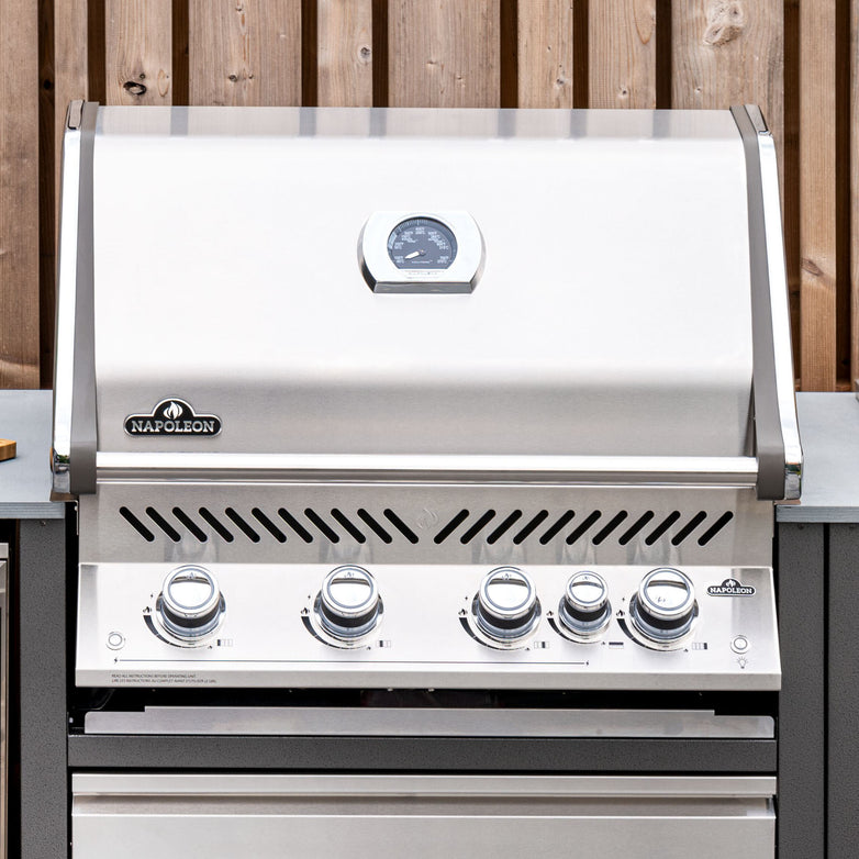A Napoleon Prestige Pro 500 Built-in Gas Grill - Natural Gas, crafted from stainless steel with four control knobs and a temperature gauge, enhanced by an infrared burner, set against a wooden fence backdrop.