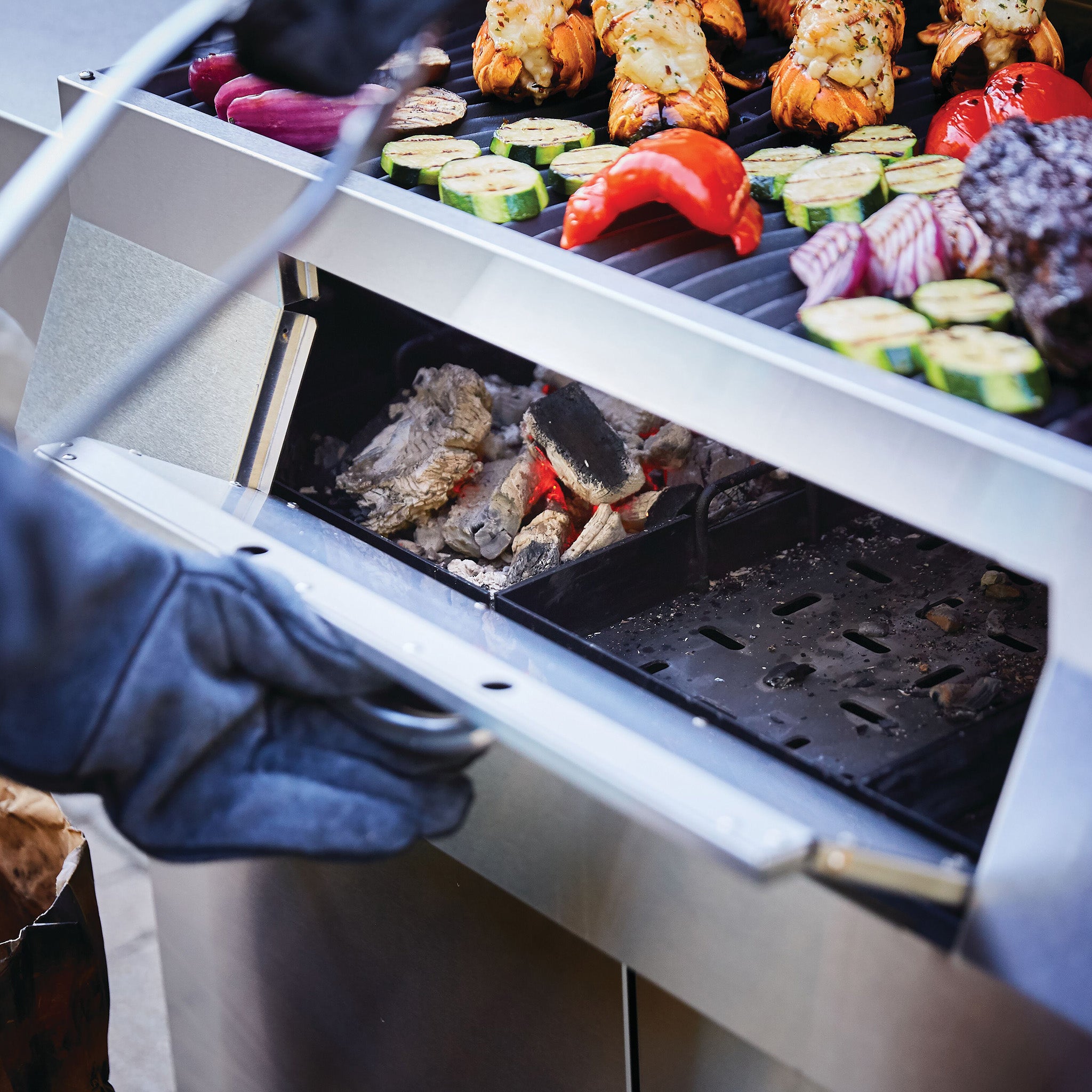 A person in gloves adjusts a Napoleon Charcoal Professional Barbecue with vegetables and lobster tails cooking on the stainless steel grate above.