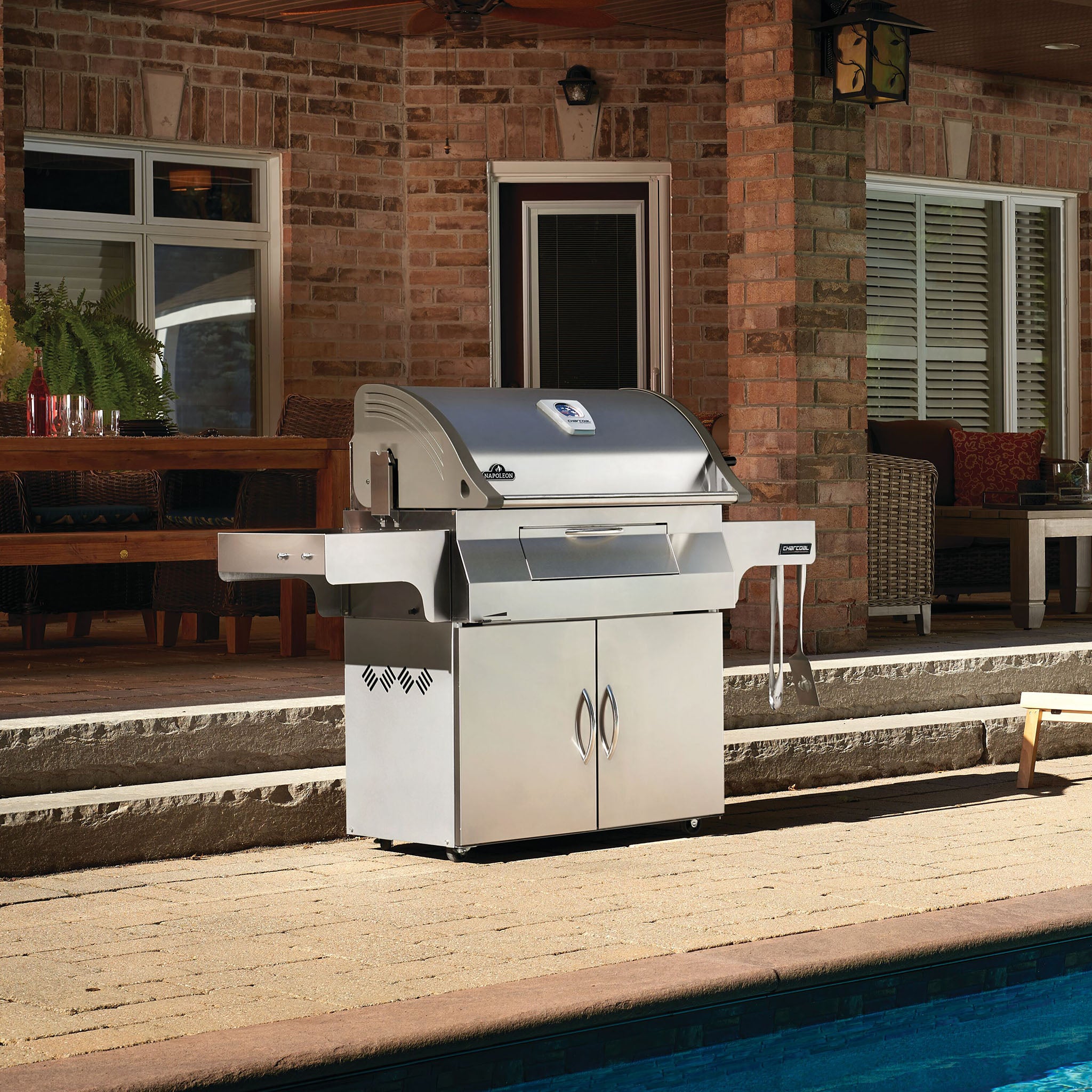 A Napoleon Charcoal Professional Barbecue sits on the patio next to a swimming pool, with a stone-faced house and patio furniture in the background.