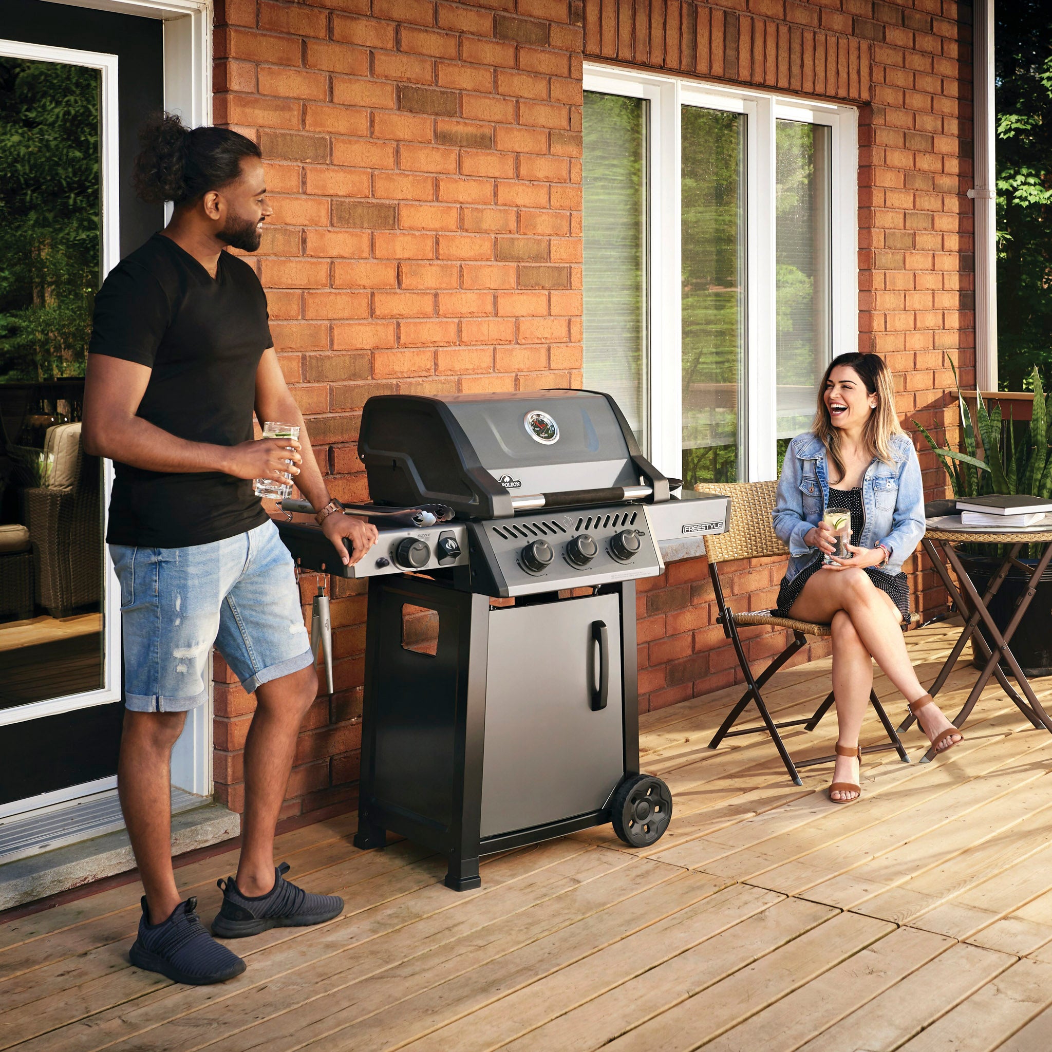A man and a woman enjoy drinks on a patio while grilling on a Napoleon Freestyle 365 Gas Barbecue with cast iron cooking grids, the JETFIRE ignition system ensuring quick starts; the man stands by the grill as the woman takes a seat.