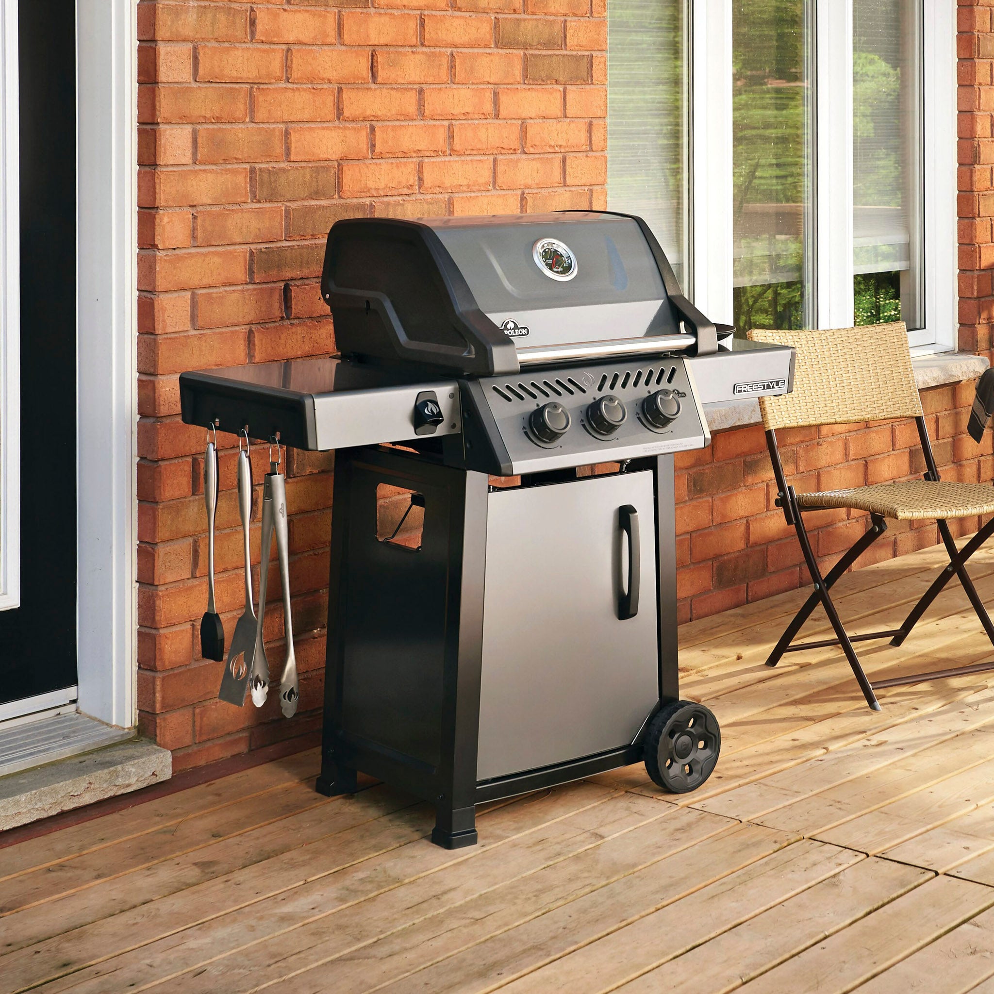 A Napoleon Freestyle 365 Gas Barbecue with a JETFIRE ignition system and cast iron cooking grids, featuring utensils hanging on the side, is placed on a wooden deck next to a brick wall and outdoor chair.