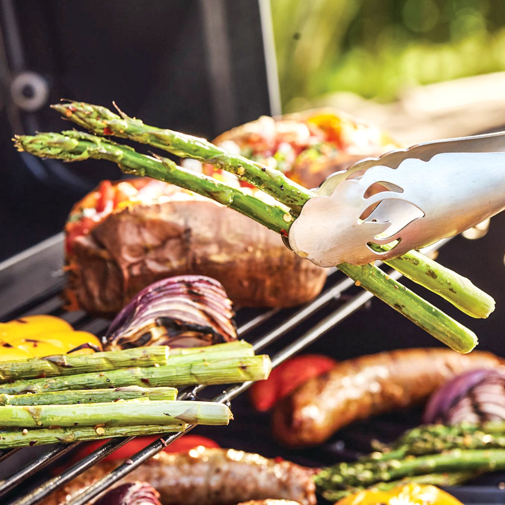 The Napoleon 3-Piece PRO Stainless Steel Grill Toolset's tongs hold grilled asparagus over a barbecue with other vegetables and sausages.