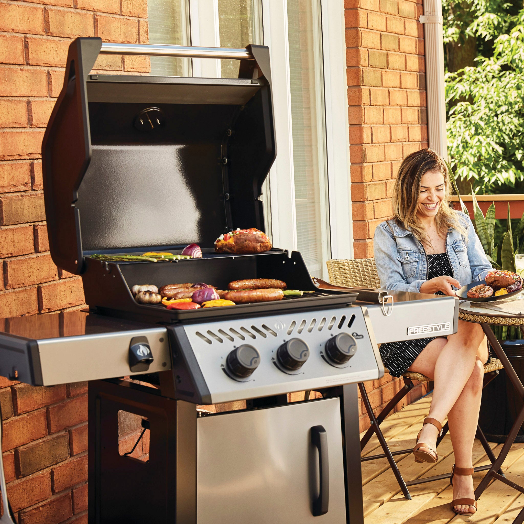 A woman enjoys a meal on a patio next to her Napoleon Freestyle 365 Gas Barbecue, savoring the aroma from the cast iron cooking grids as they cook various foods, including vegetables and meat. The JETFIRE ignition system ensures everything cooks perfectly.